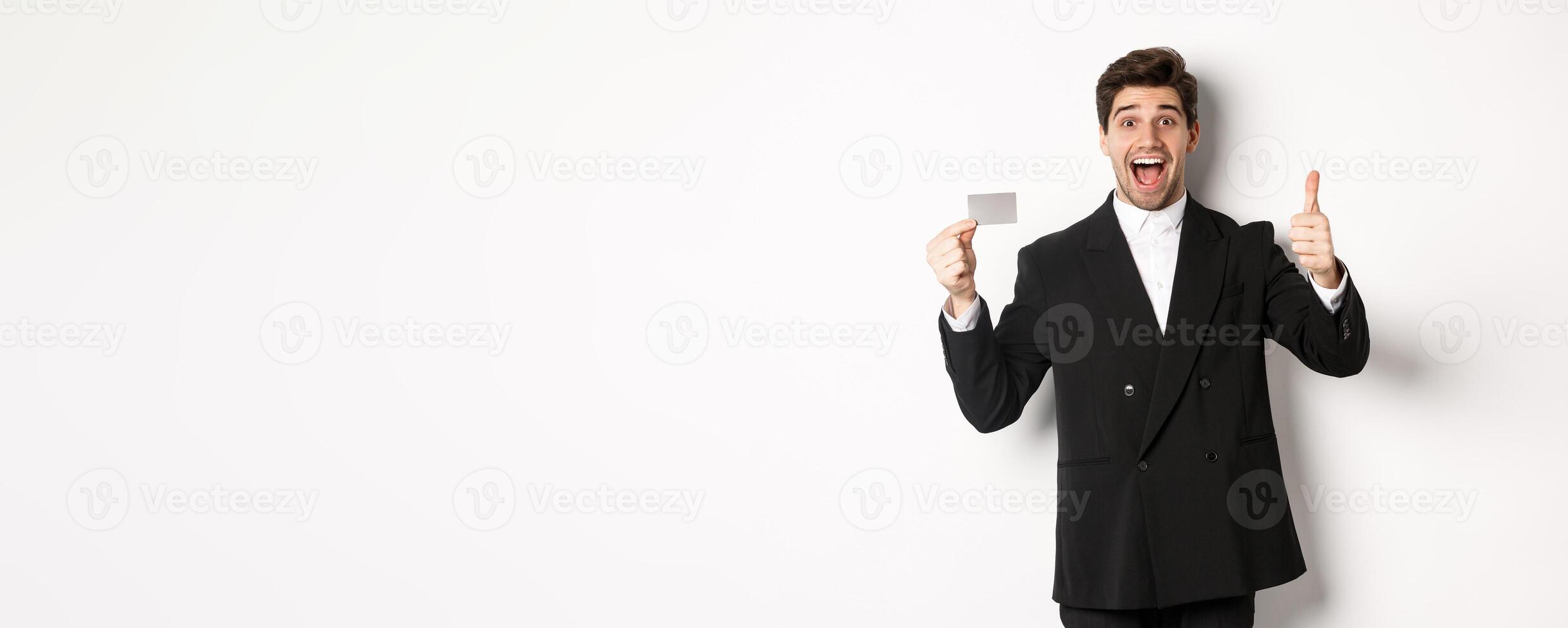 Image of good-looking businessman in black suit, showing thumbs-up and credit card, recommending bank, standing against white background photo