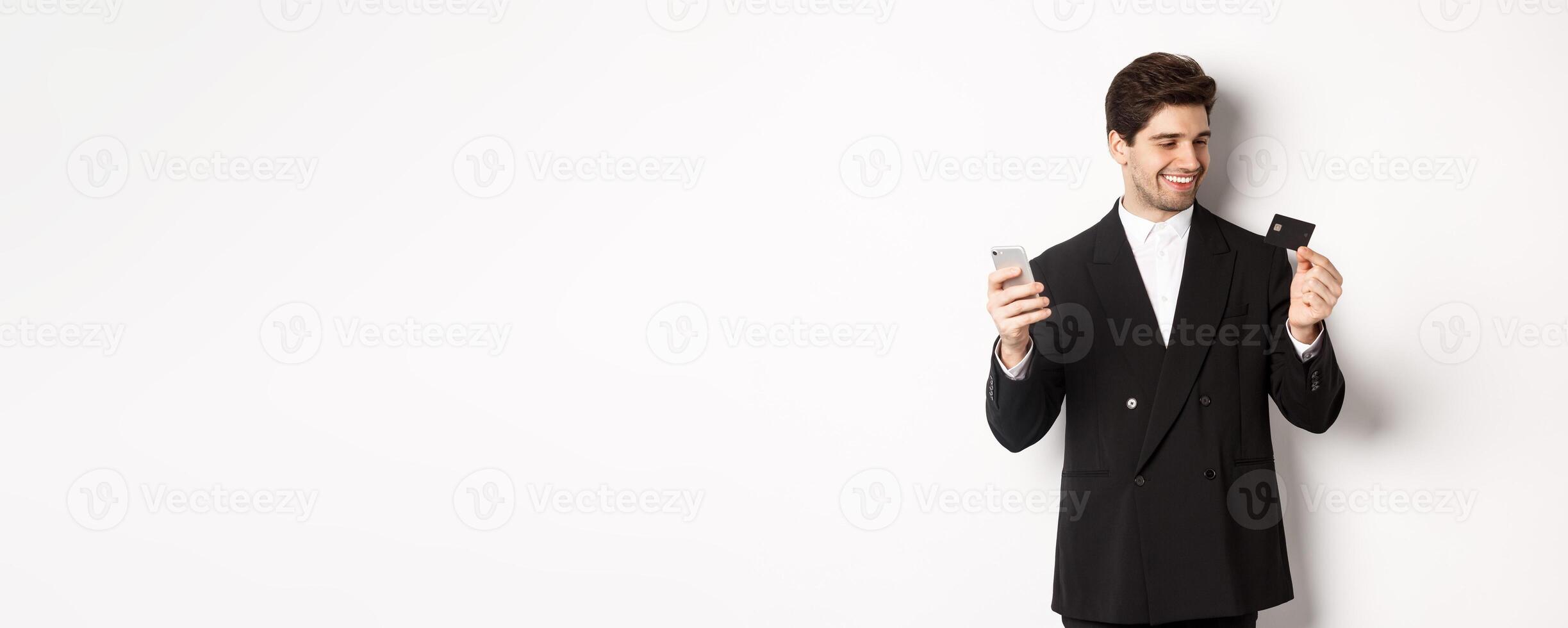 Portrait of confident handsome businessman shopping online, holding credit card and smartphone, standing over white background photo