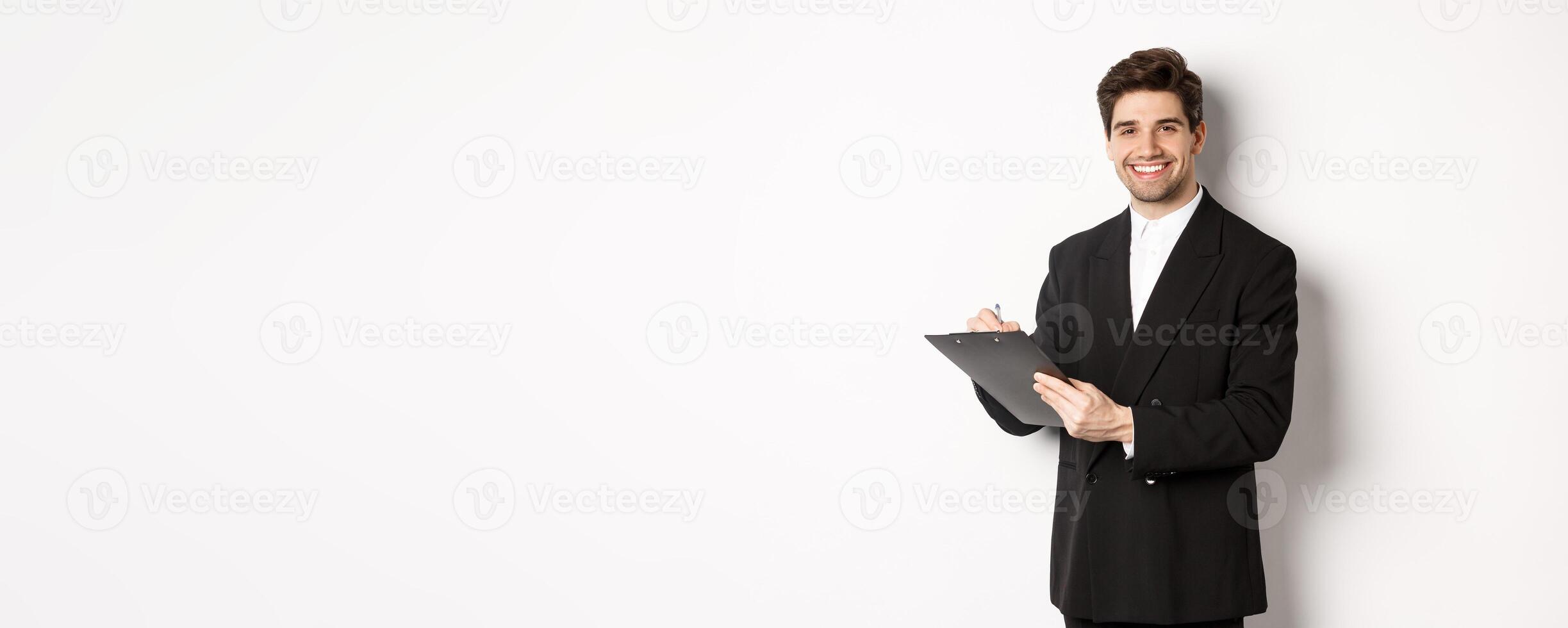 Portrait of confident businessman in black suit, signing documents and smiling, standing happy against white background photo