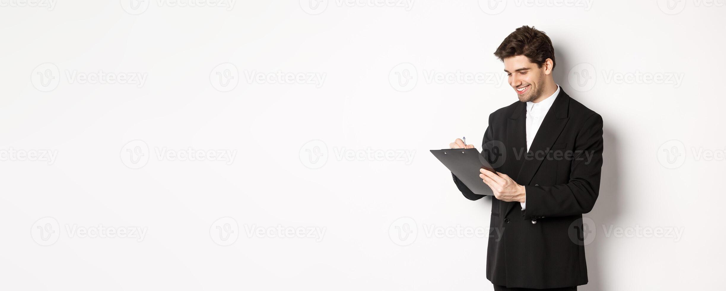 Image of handsome businessman in suit signing documents, looking at clipboard and smiling, standing against white background photo