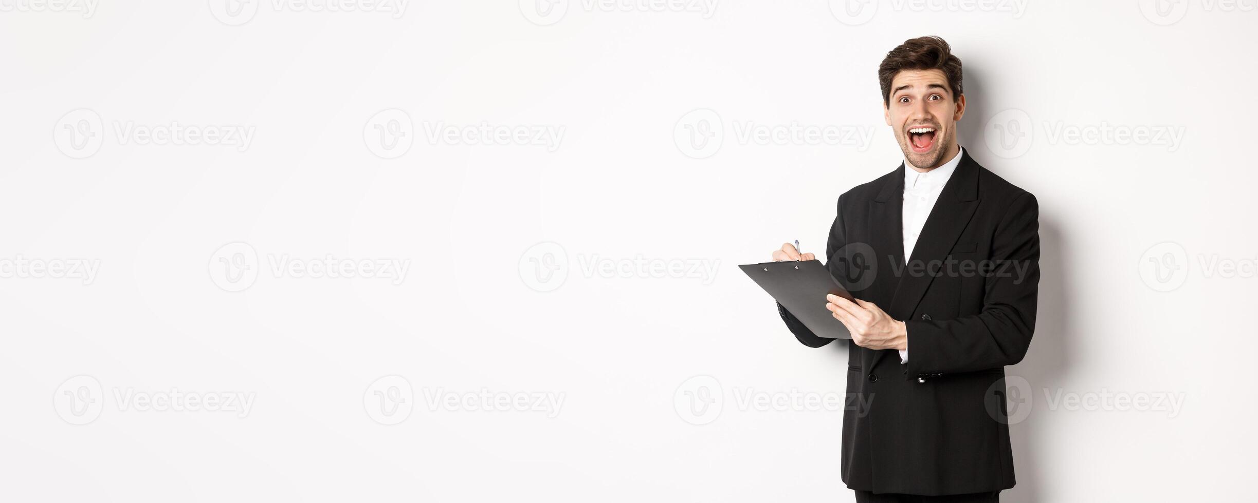 Portrait of handsome businessman in black suit, looking amazed while signing documents on clipboard, standing against white background photo