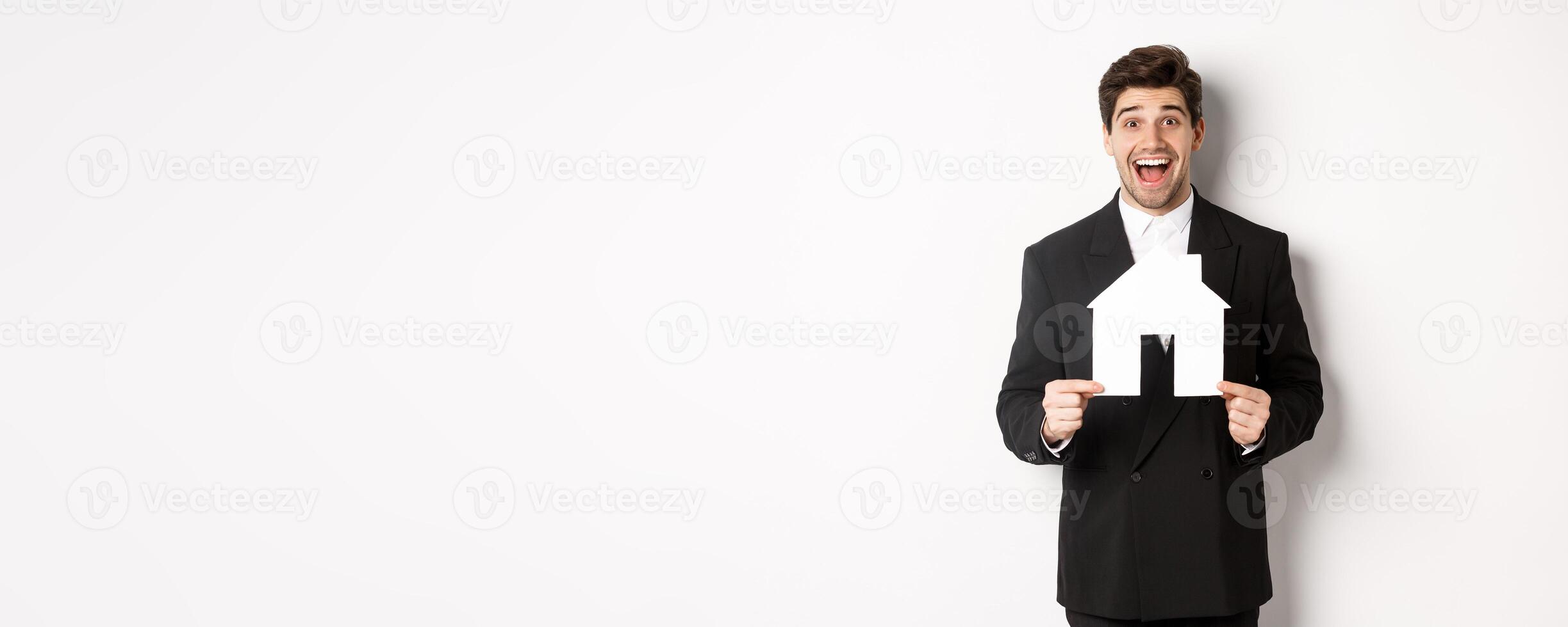 Image of handsome real estate agent in black suit showing home maket, and looking amazed, selling houses, standing against white background photo
