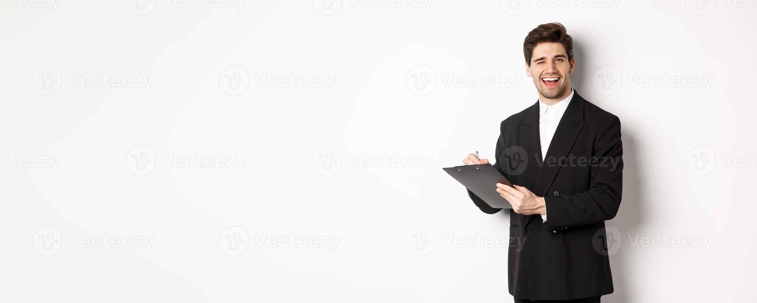 Image of handsome, successful boss in black suit, winking and smiling while signing documents, standing against white background photo