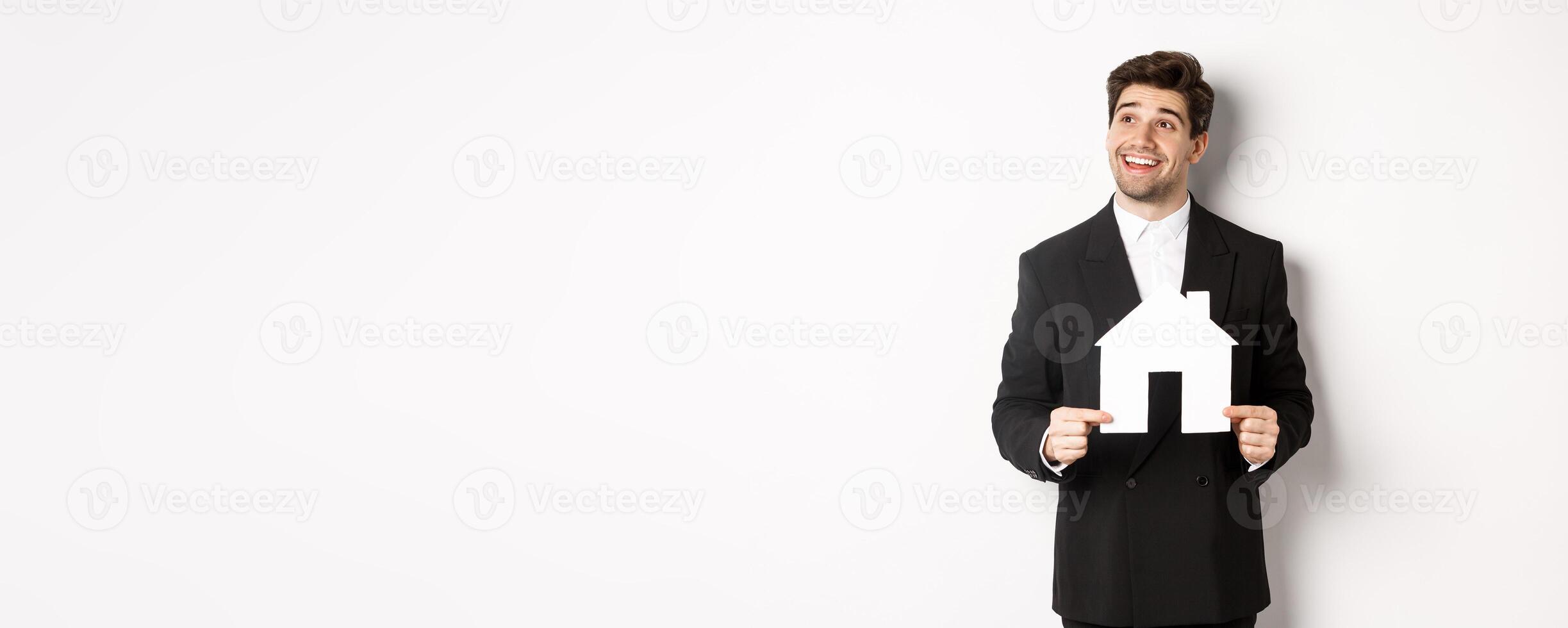 Image of handsome investor in black suit, holding house maket and looking at upper left corner thoughtful, standing against white background photo