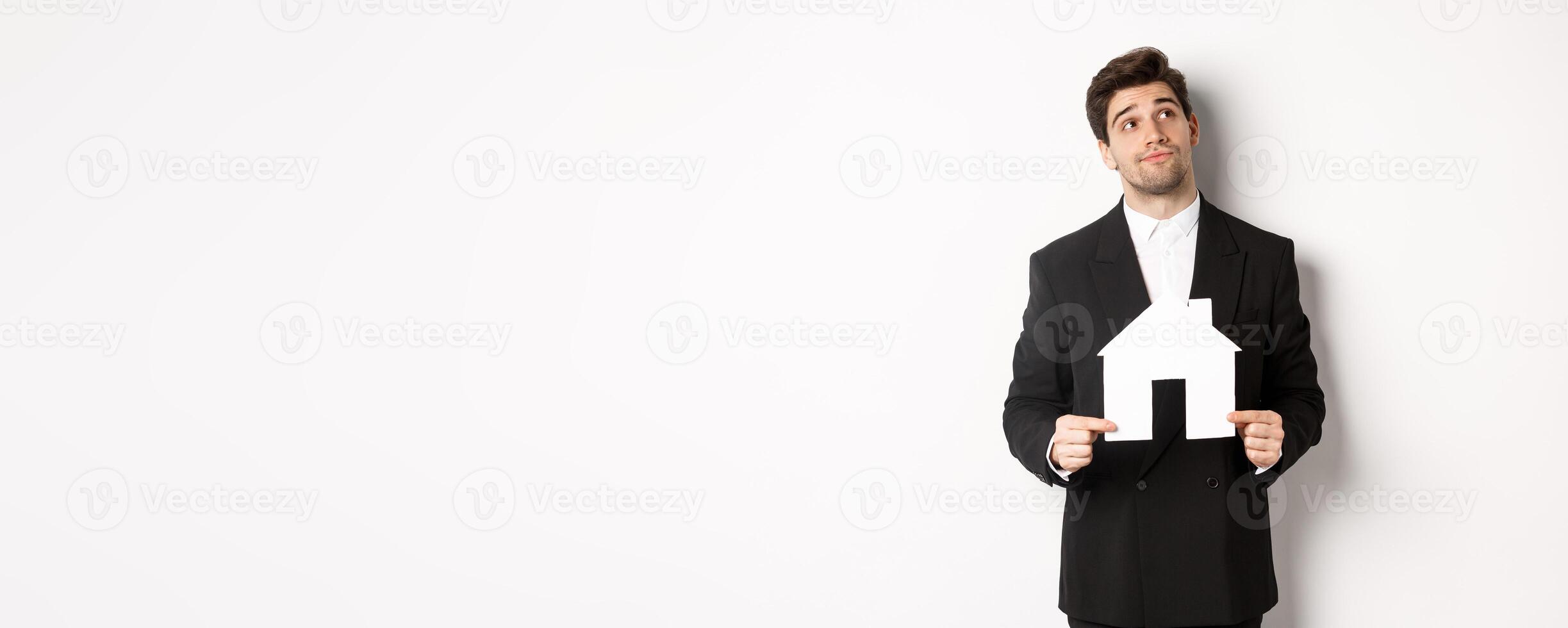 Image of handsome businessman in black suit, looking for home, holding house maket and gazing dreamy at upper right corner, standing against white background photo