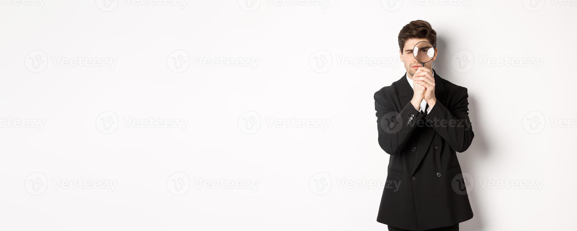 Image of serious businessman in black trendy suit, looking through magnifying glass, looking for employees, standing against white background photo