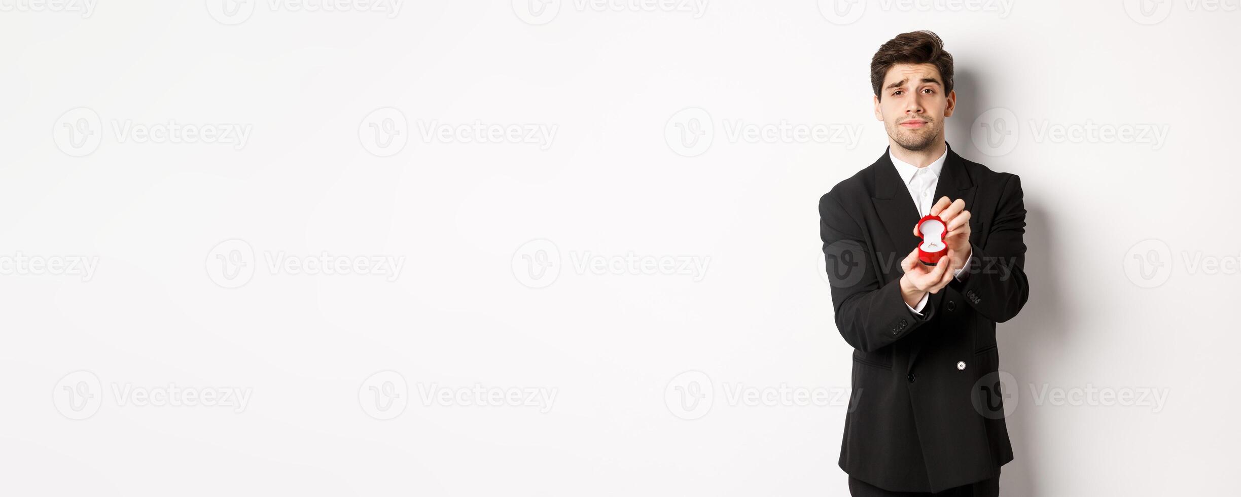 Portrait of handsome man making a proposal, asking to marry him, showing ring, standing in black suit against white background photo