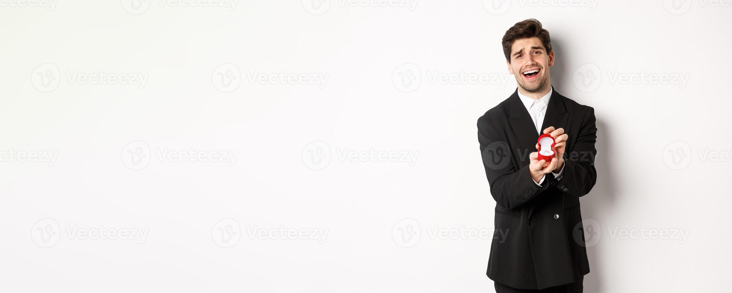 Portrait of handsome man in black suit, open box with wedding ring, making a proposal, asking to marry him, standing against white background photo