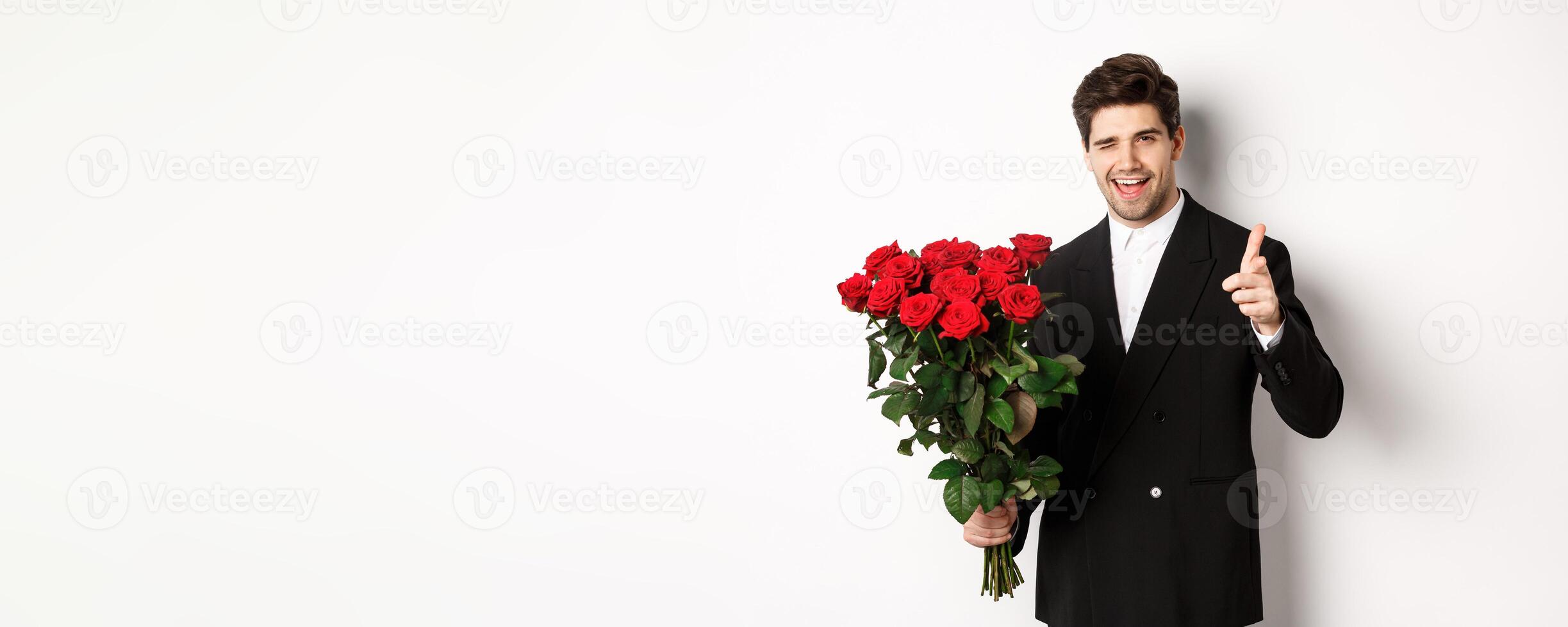 Image of handsome romantic guy in black suit, holding bouquet of roses and pointing at camera, congratulating with holiday, standing against white background photo