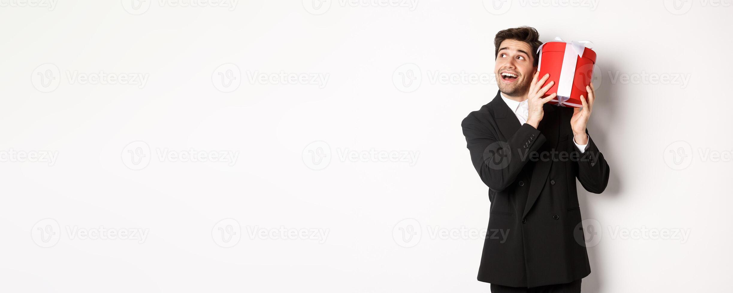 Concept of christmas holidays, celebration and lifestyle. Image of excited man enjoying new year, shaking gift box to guess what inside, standing against white background photo