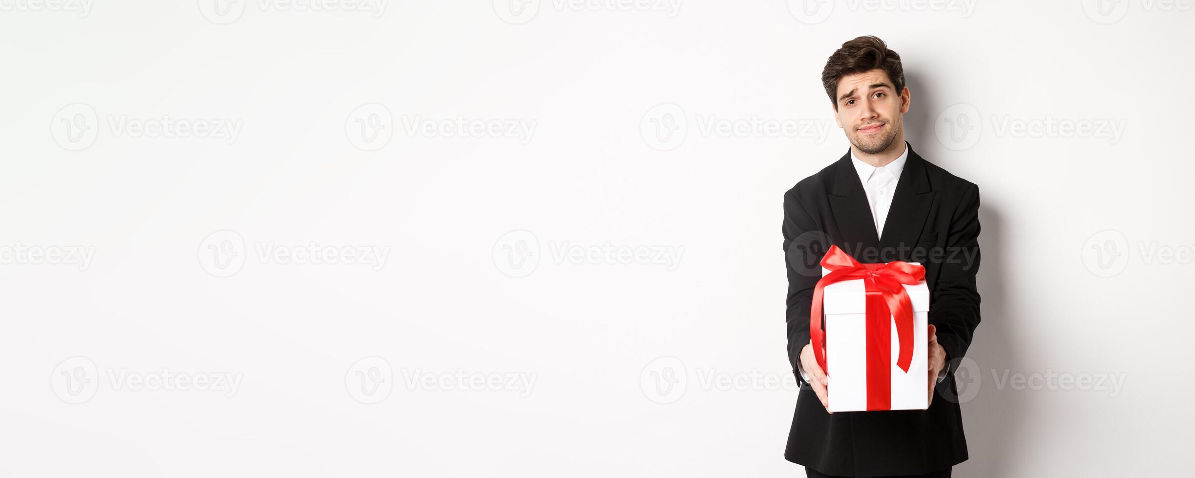 Concept of christmas holidays, celebration and lifestyle. Image of handsome boyfriend in black suit, giving you a present, extending hands with gift, standing over white background photo
