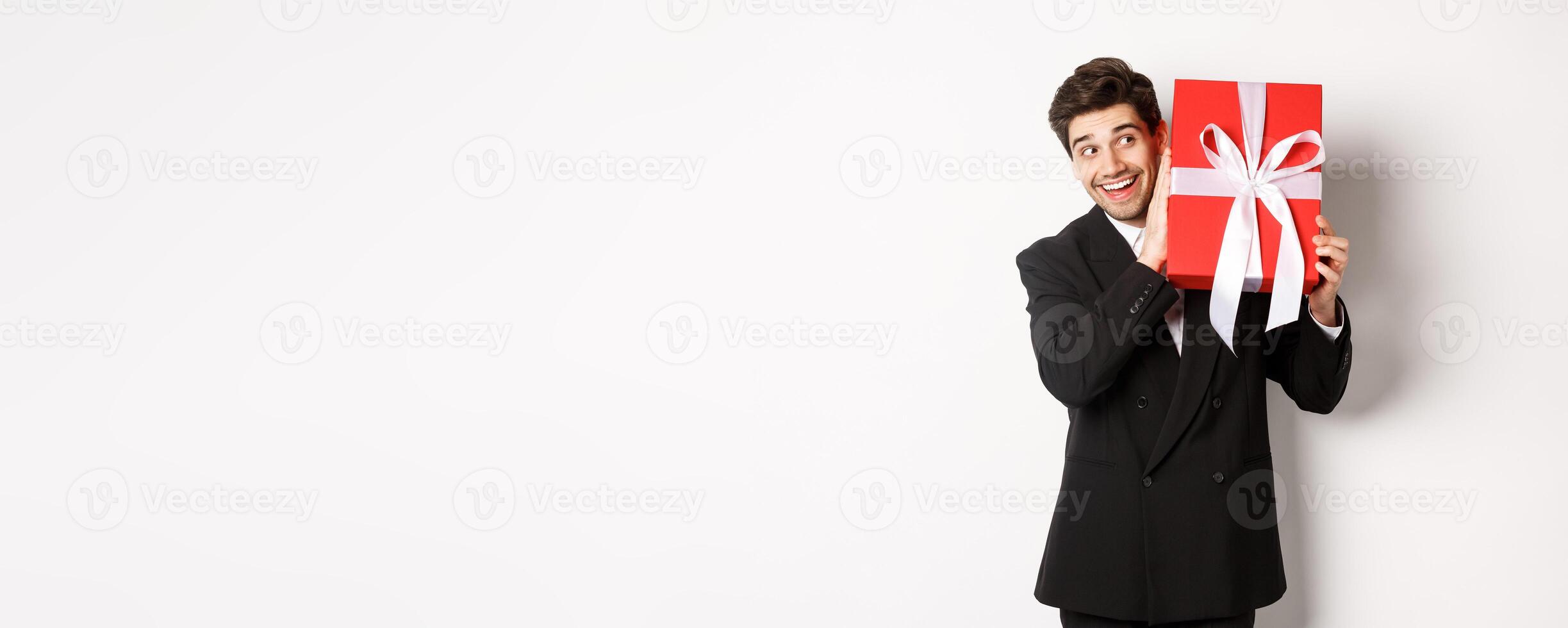 Concept of christmas holidays, celebration and lifestyle. Image of excited man enjoying new year, shaking gift box to guess what inside, standing against white background photo