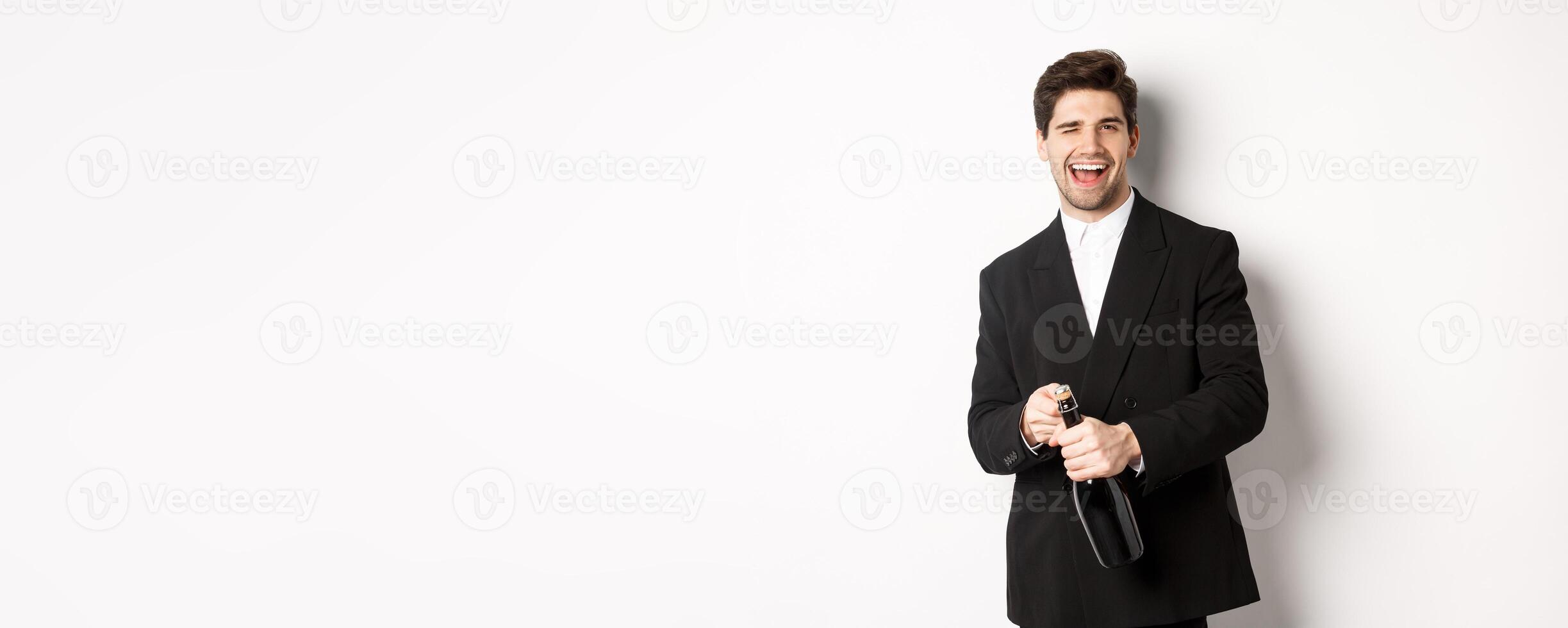 Image of handsome, confident man in black suit, celebrating holiday, open a bottle of champagne and partying, standing joyful against white background photo