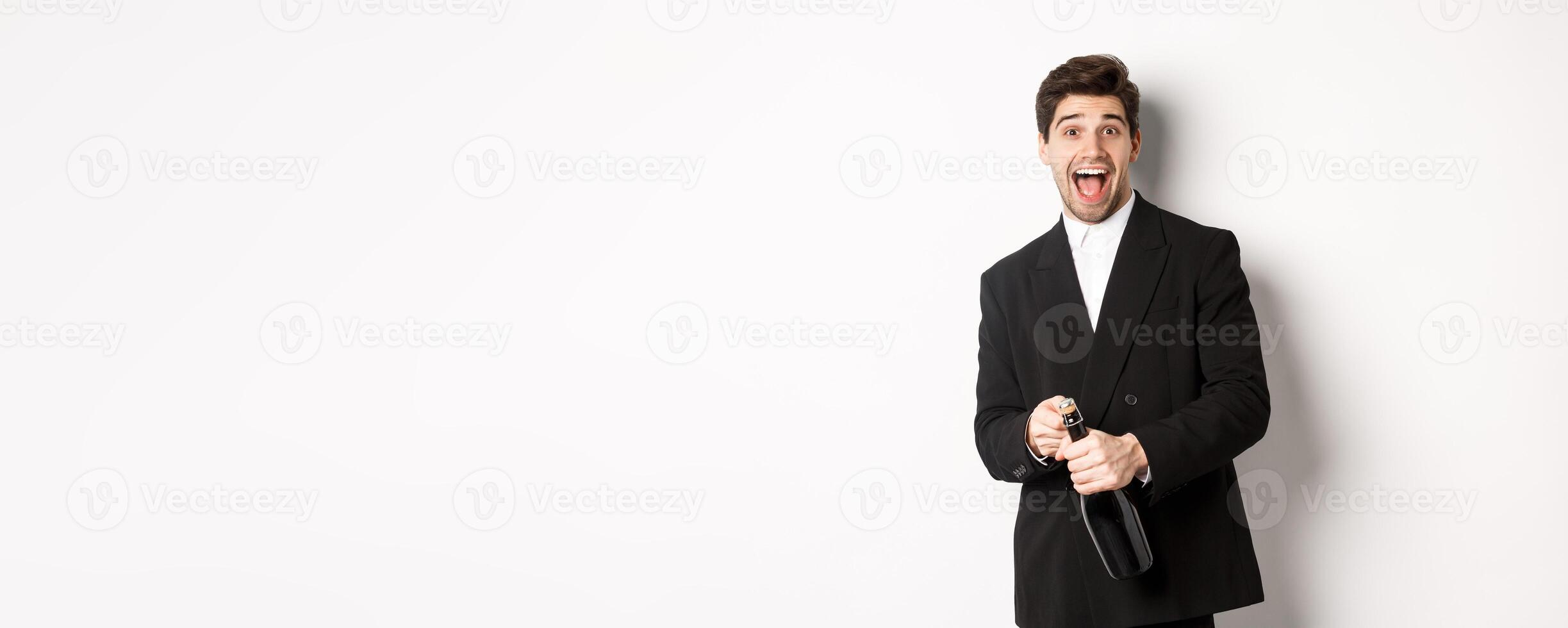 Image of attractive man in black suit having a party, celebrating new year and opening bottle of champagne, standing happy against white background photo