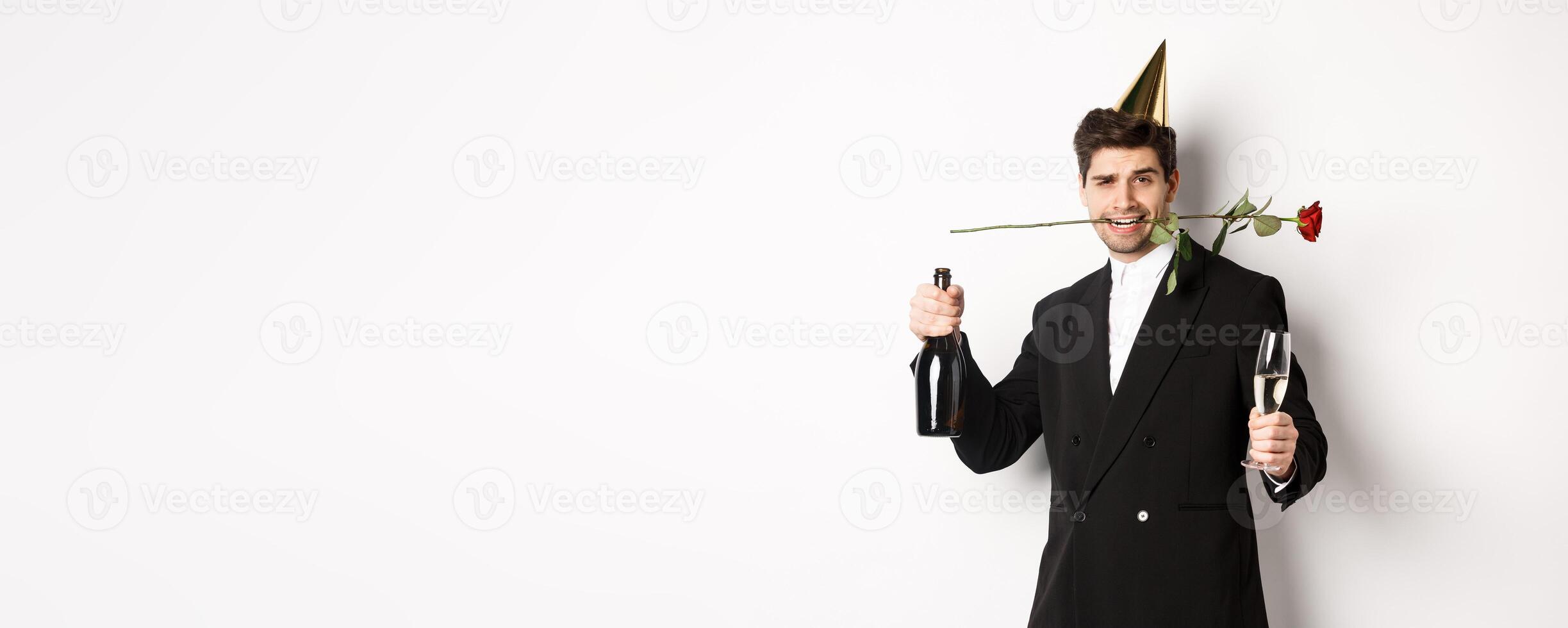 Romantic guy in trendy suit, celebrating and having a party, holding rose in teeth and champagne, standing over white background photo