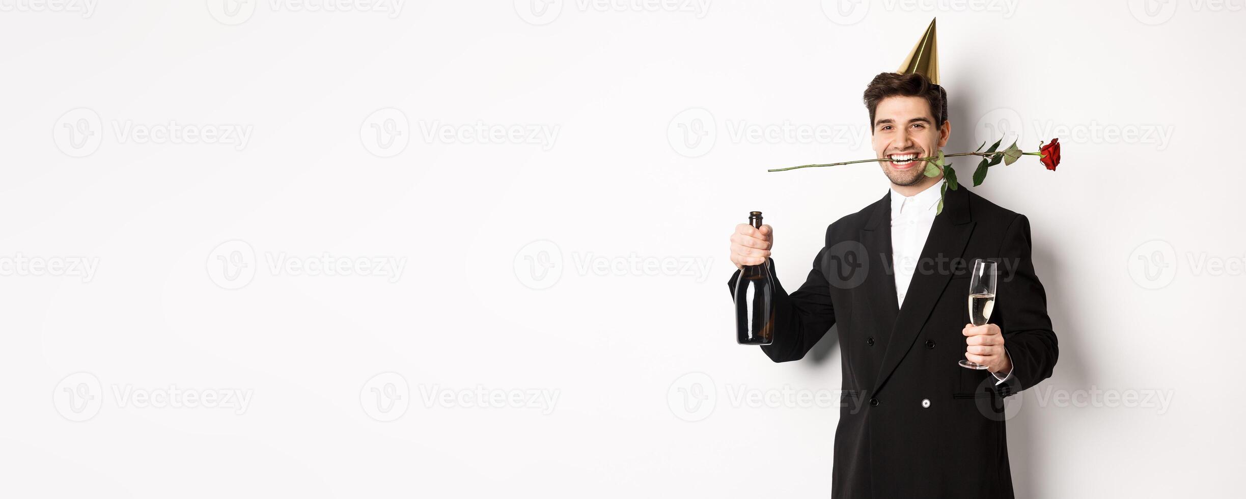 Funny guy in trendy suit, celebrating and having a party, holding rose in teeth and champagne, standing over white background photo