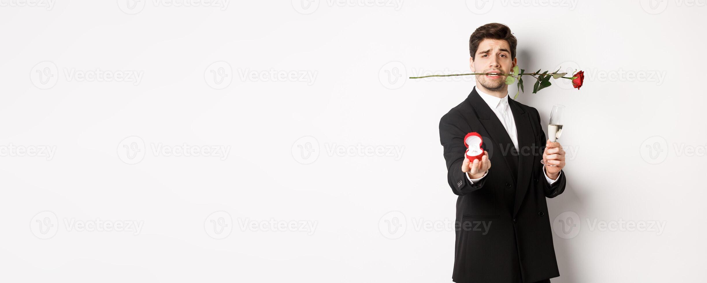 Romantic young man in suit making a proposal, holding rose in teeth and glass of champagne, showing engagement ring, asking to marry him, standing against white background photo