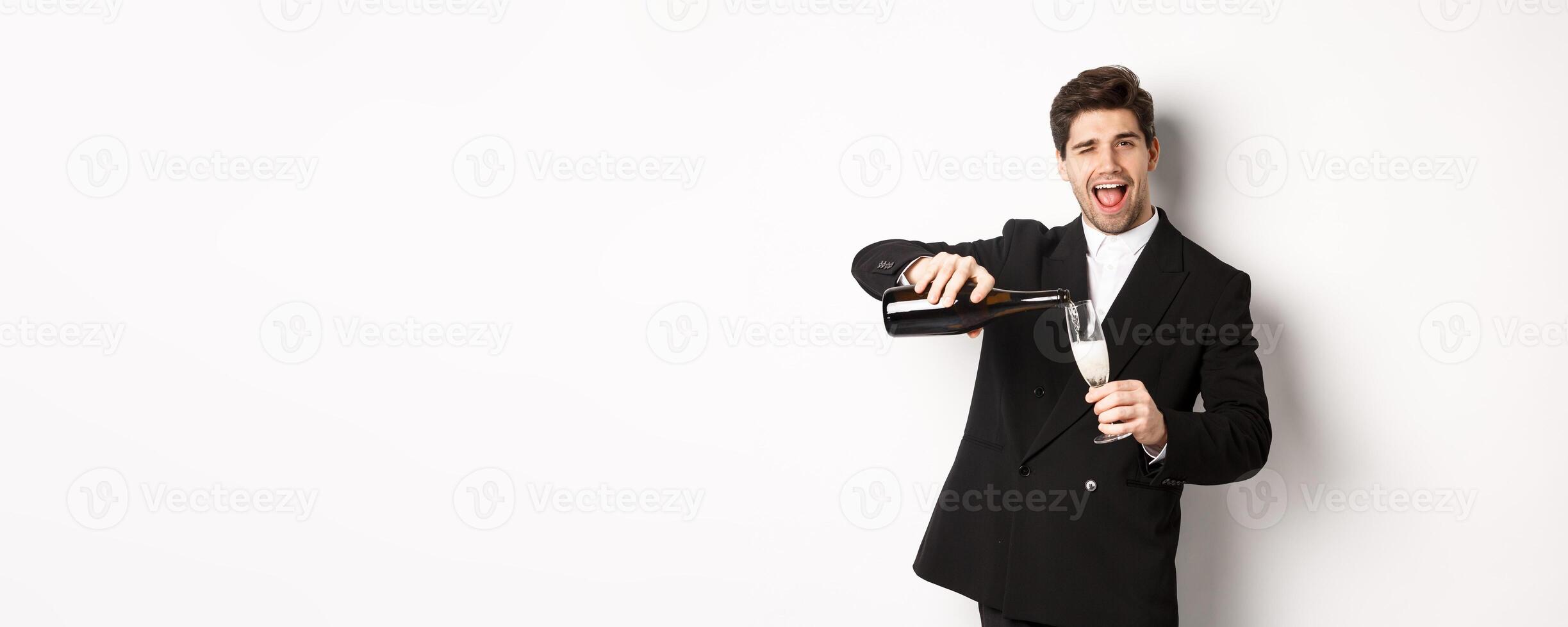 Handsome man in trendy suit pouring glass of champagne, celebrating christmas, smiling amazed and having fun, standing against white background photo