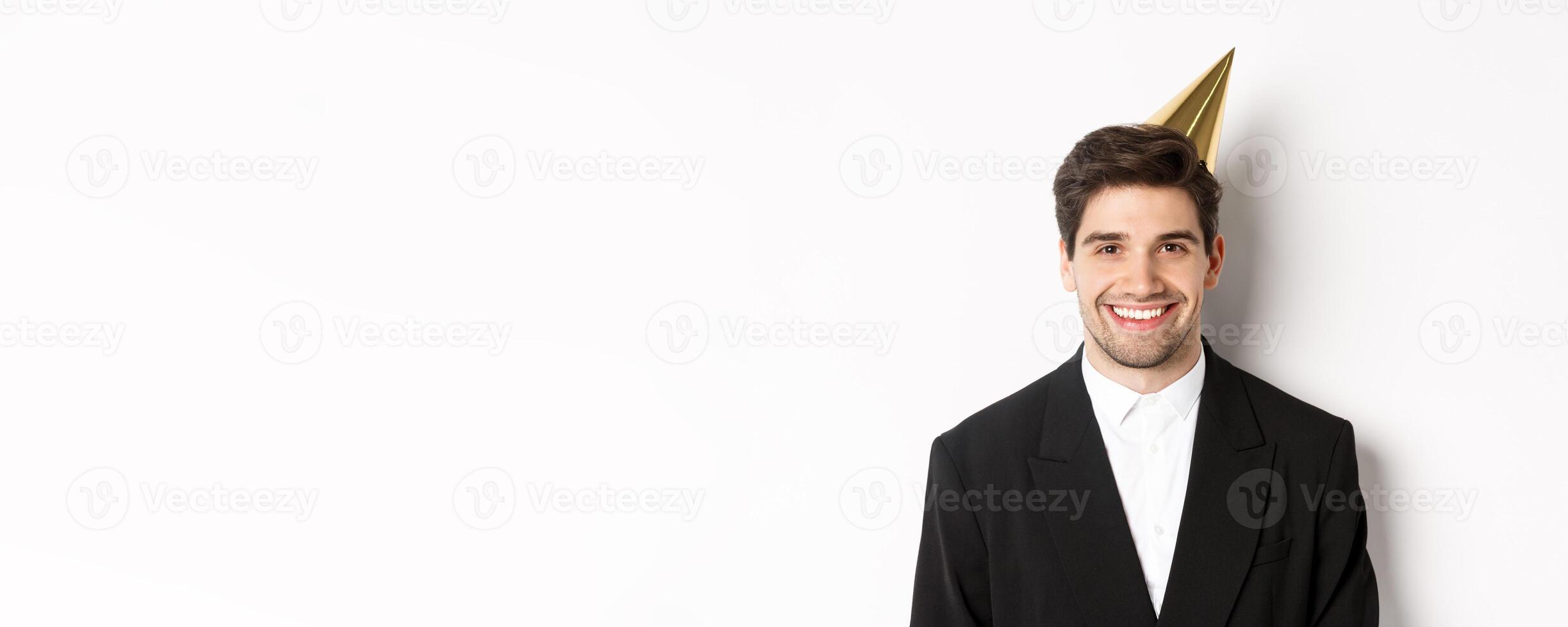 Close-up of handsome happy man in suit and party hat, smiling joyful, celebrating holiday, standing against white background photo