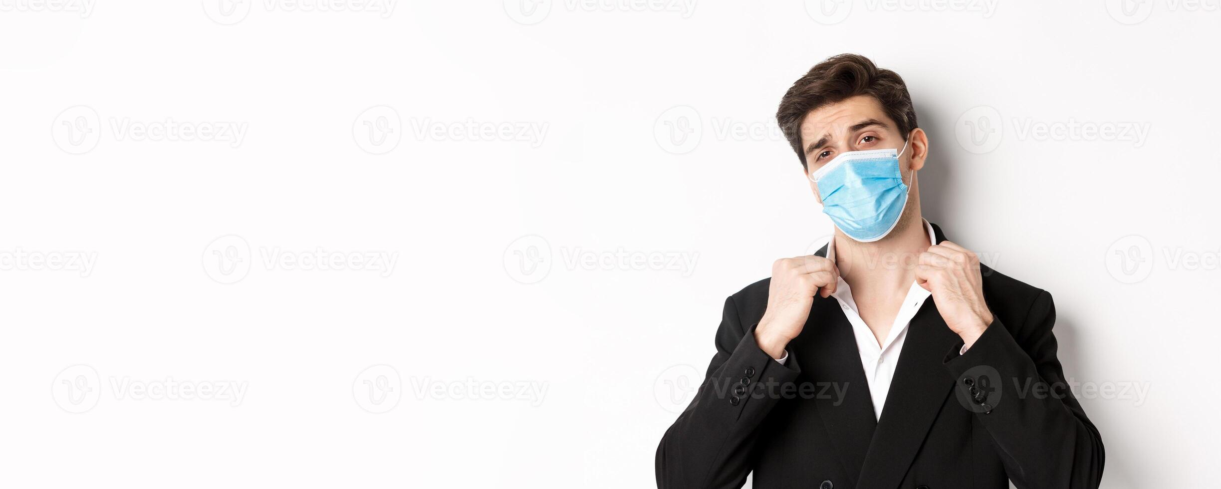 Concept of covid-19, business and social distancing. Image of confident handsome man in trendy suit and medical mask, looking sassy, standing against white background photo