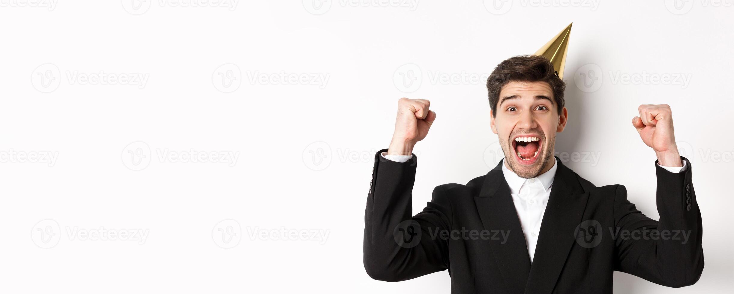 Close-up of happy good-looking man, wearing party hat and suit, raising hands up and rejoicing, celebrating new year, standing against white background photo