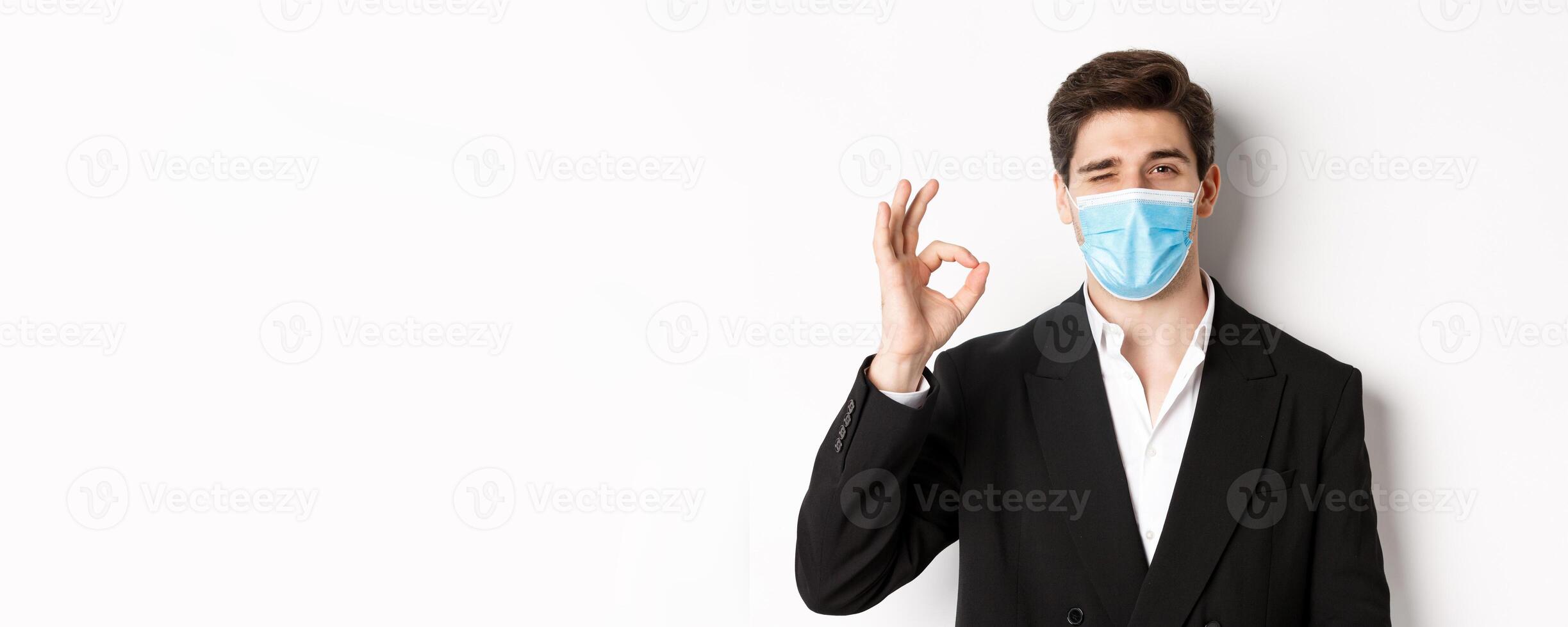 Concept of covid-19, business and social distancing. Close-up of good-looking businessman in trendy suit and medical mask, showing okay sign and winking, standing against white background photo