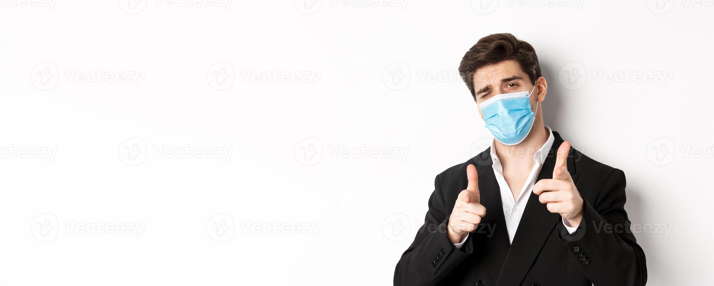Concept of covid-19, business and social distancing. Close-up of sassy man in trendy suit and medical mask, pointing fingers at camera, standing against white background photo