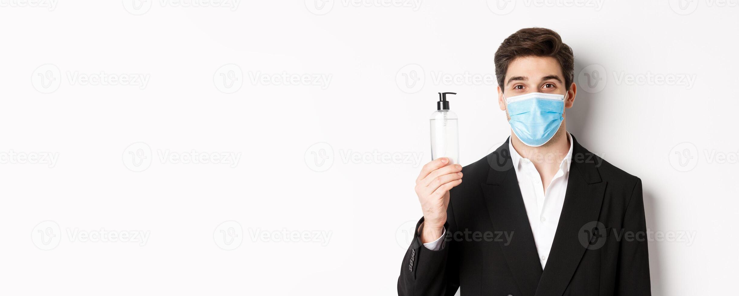 Concept of covid-19, business and social distancing. Close-up of handsome man in trendy suit and medical mask, showing hand sanitizer, standing against white background photo