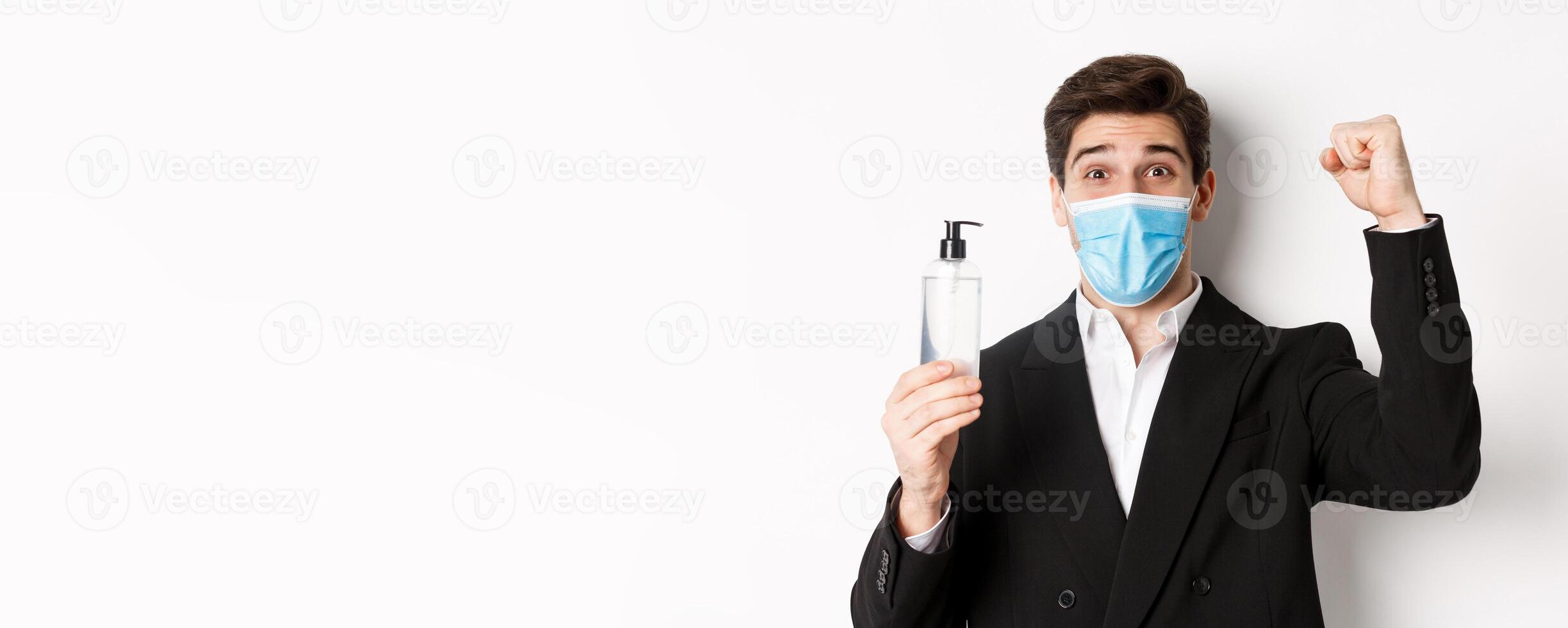 Concept of covid-19, business and social distancing. Close-up of happy man in trendy suit and medical mask, cheering and raising hand up, showing hand sanitizer, white background photo