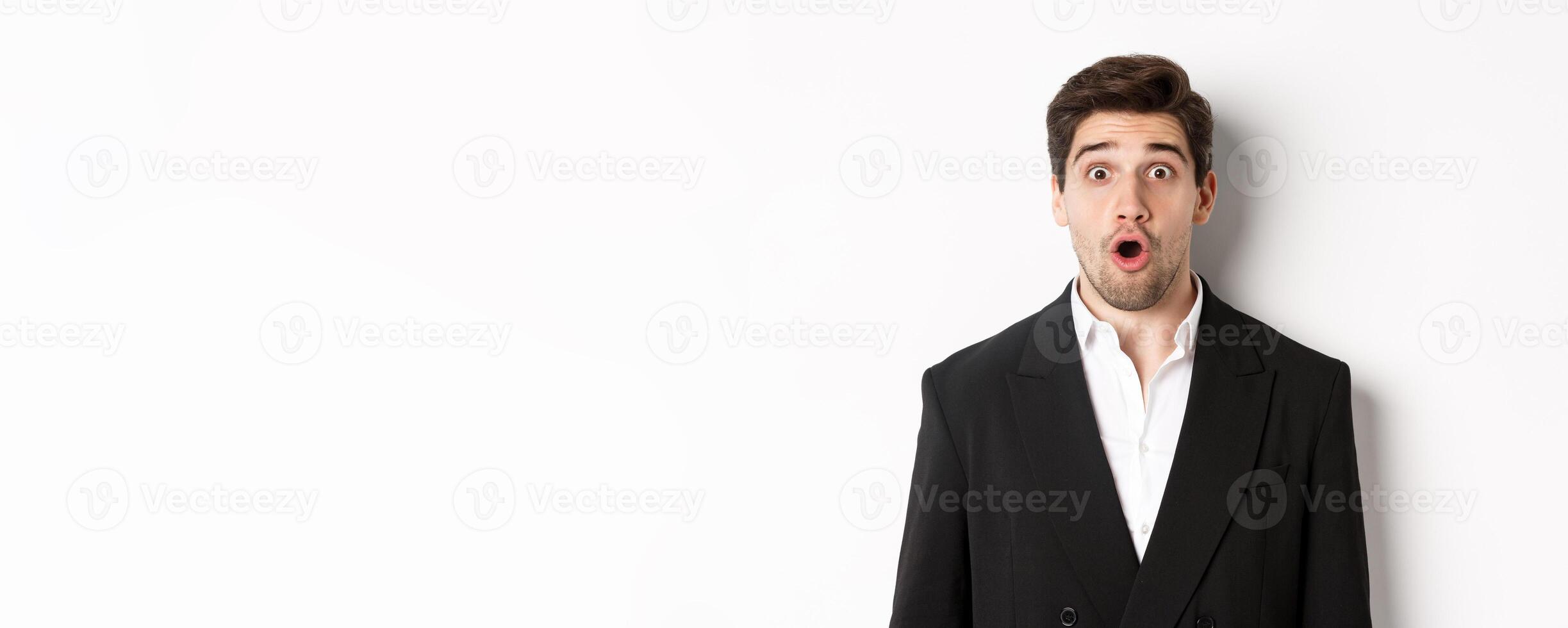 Close-up of attractive man in black suit, looking surprised and impressed at advertisement, standing over white background photo