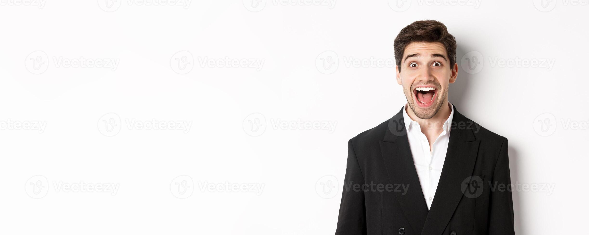 Close-up of attractive man in black suit, smiling amazed and looking at advertisement, standing over white background photo