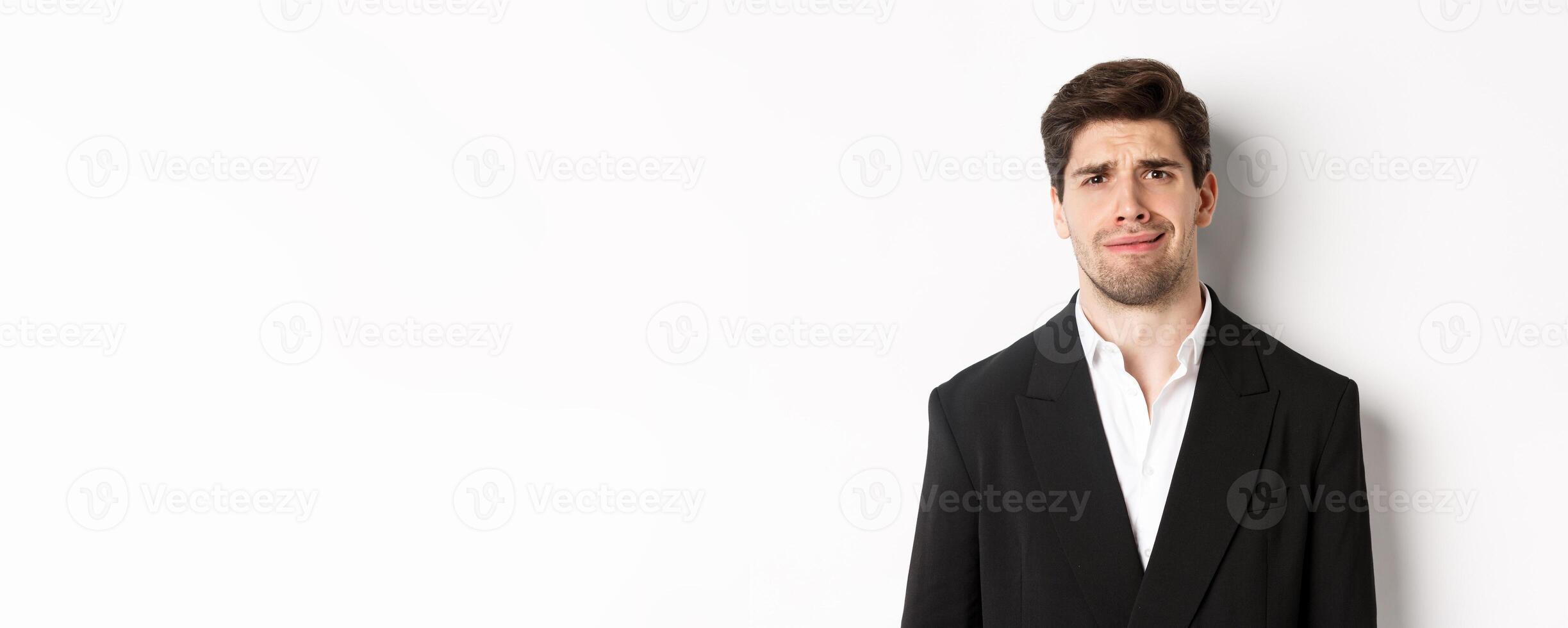 Close-up of confused handsome man in suit, grimacing and looking perplexed, wearing trendy suit, standing against white background photo