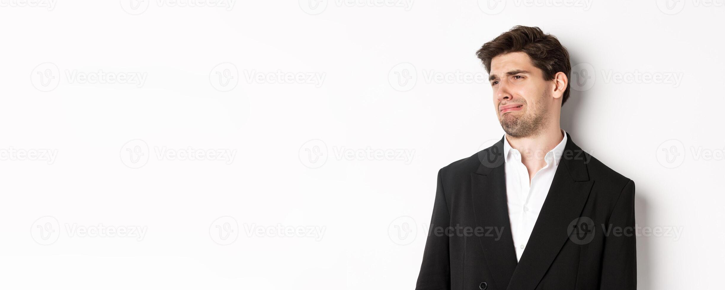 Close-up of disgusted young man in trendy suit, grimacing upset, looking left and standing against white background photo