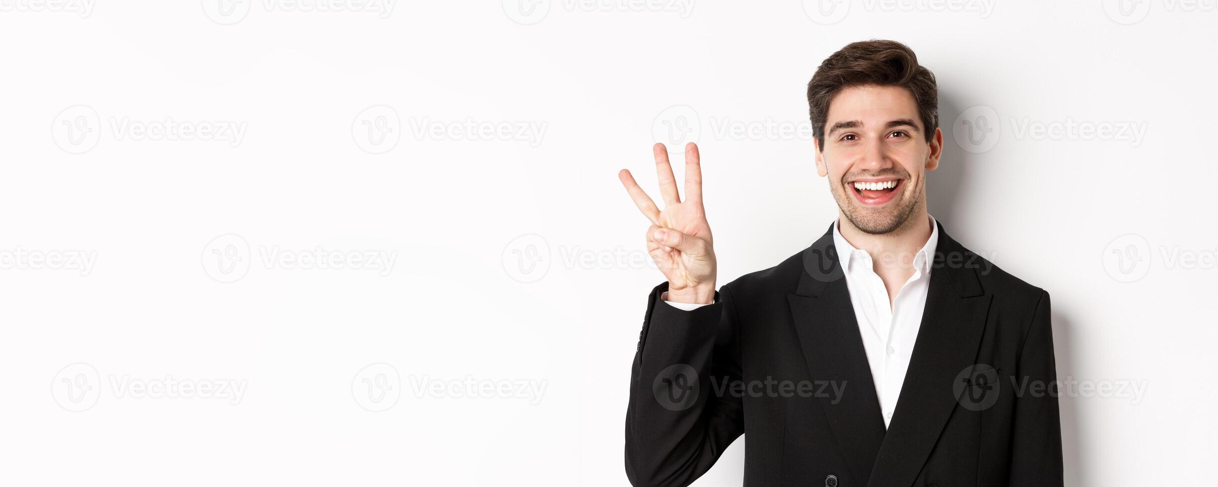 Close-up of handsome businessman in black suit, smiling amazed, showing number three, standing over white background photo