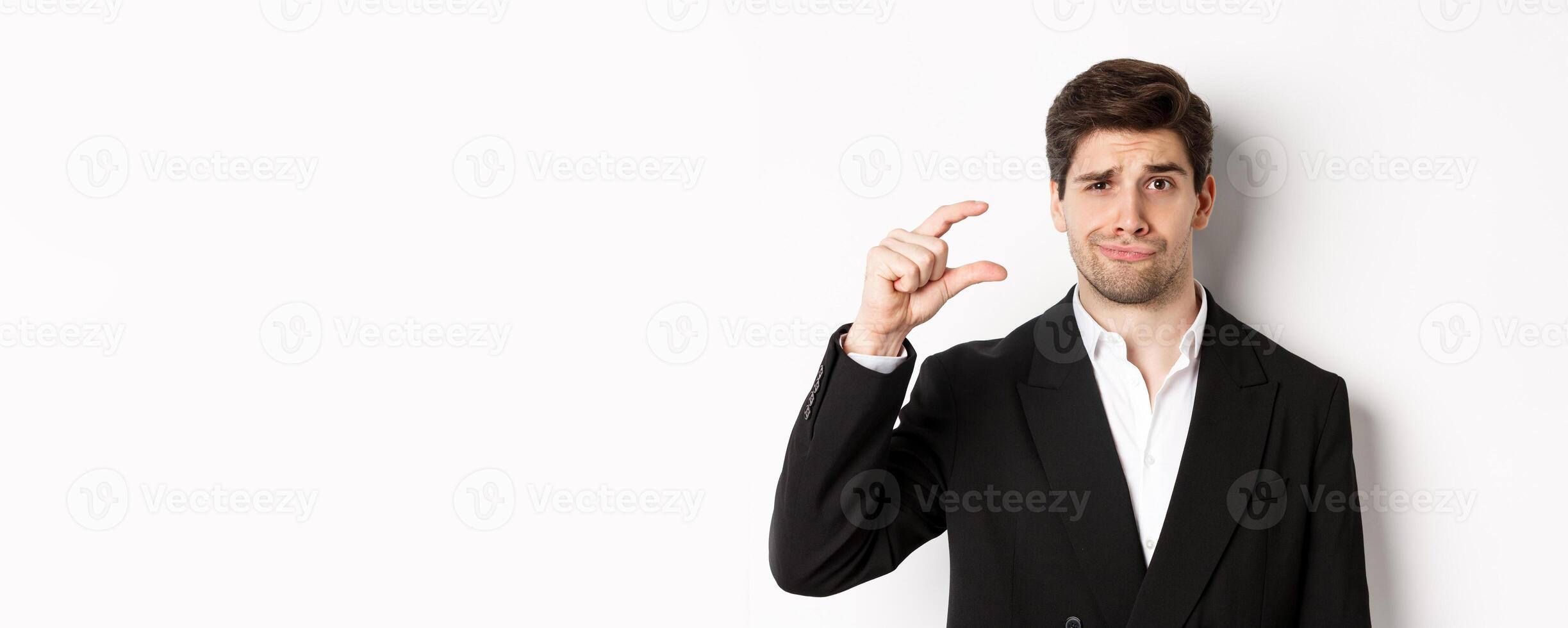 Close-up of handsome businessman in trendy suit, showing something small and looking disappointed, standing against white background photo