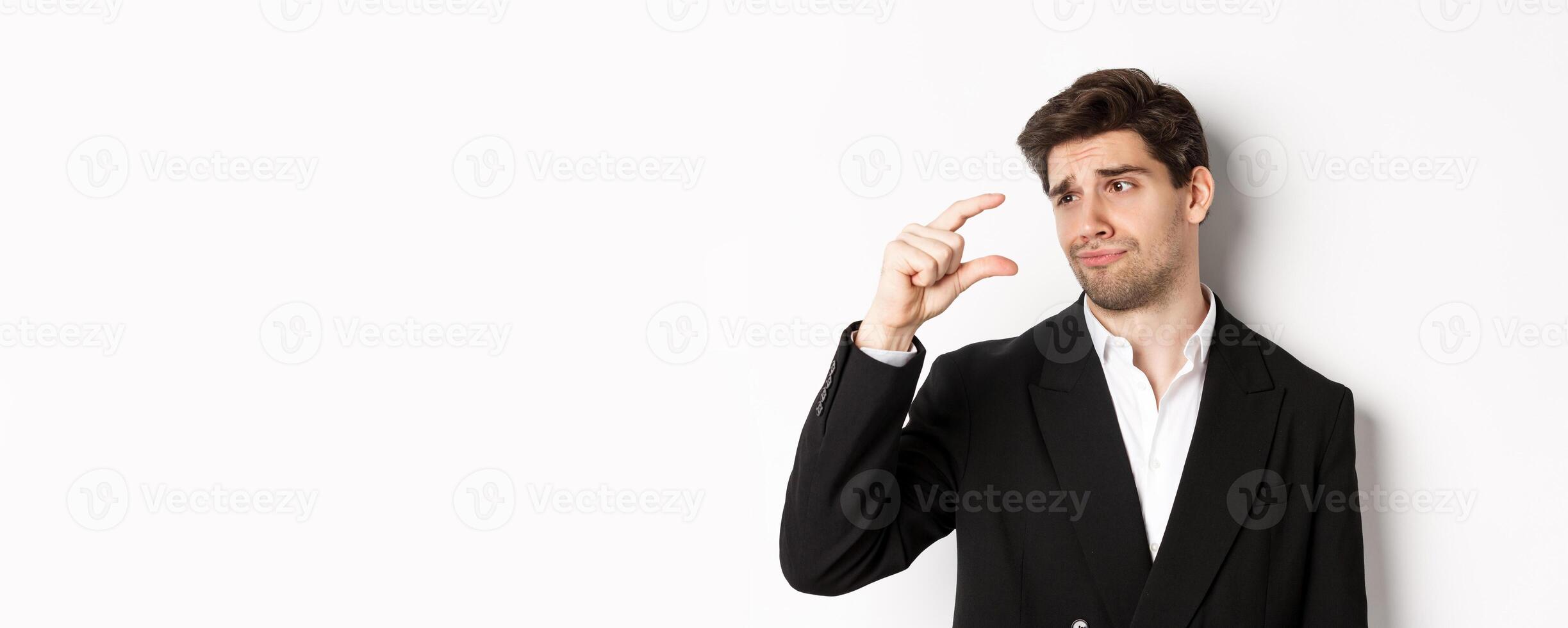 Close-up of handsome businessman in trendy suit, showing something small with disappointment, standing against white background photo
