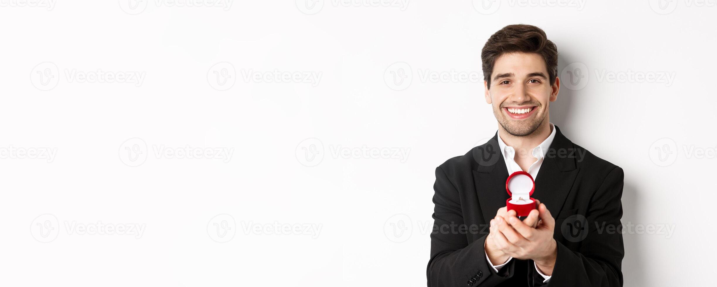 Image of handsome man looking romantic, open small box with engagement ring, making a proposal and smiling, standing against white background photo