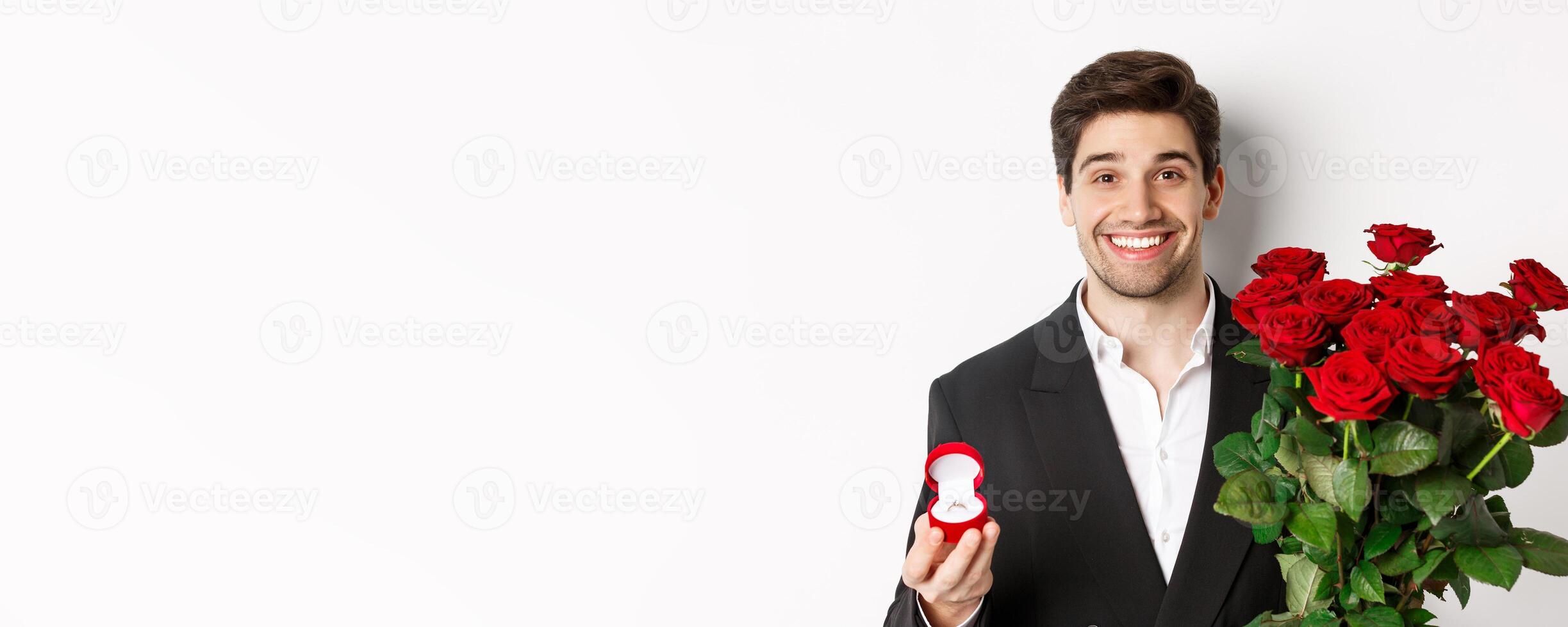 Close-up of attractive man in suit, holding bouquet of roses and engagement ring, making proposal, standing against white background photo