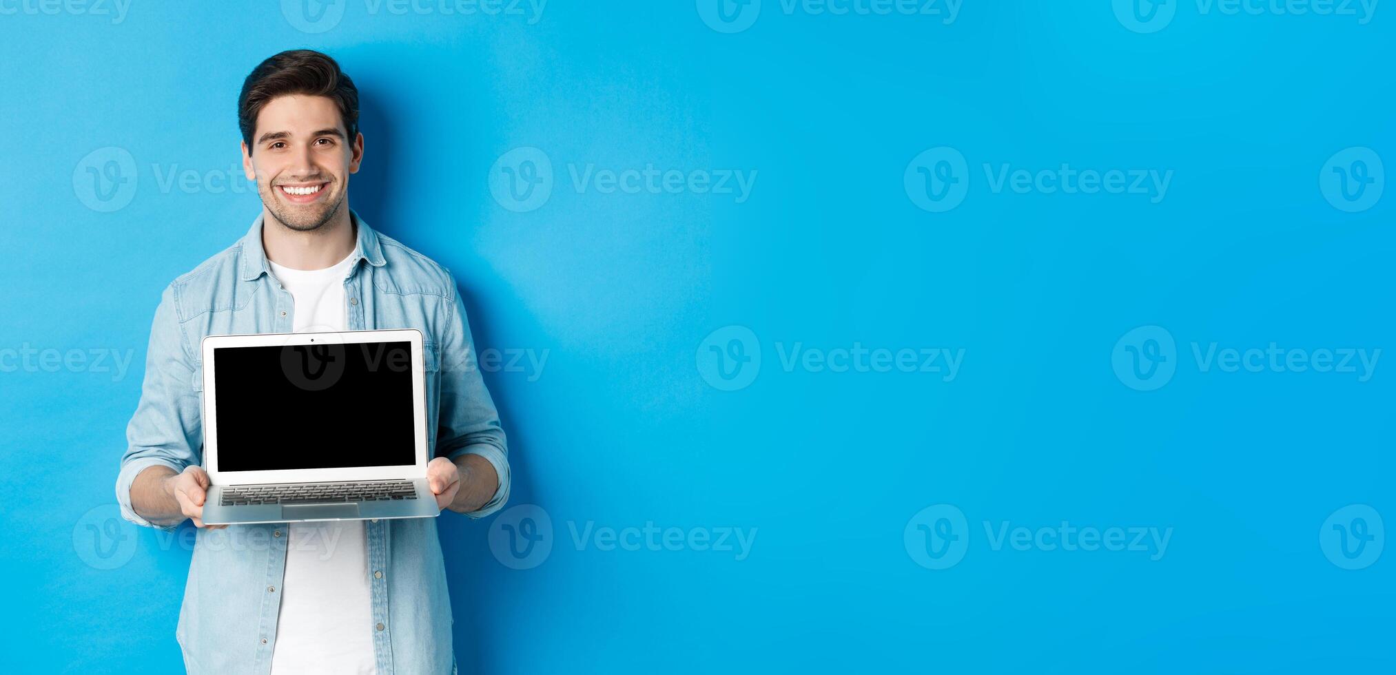 Handsome young man introduce product on laptop screen, showing computer and smiling, standing over blue background photo