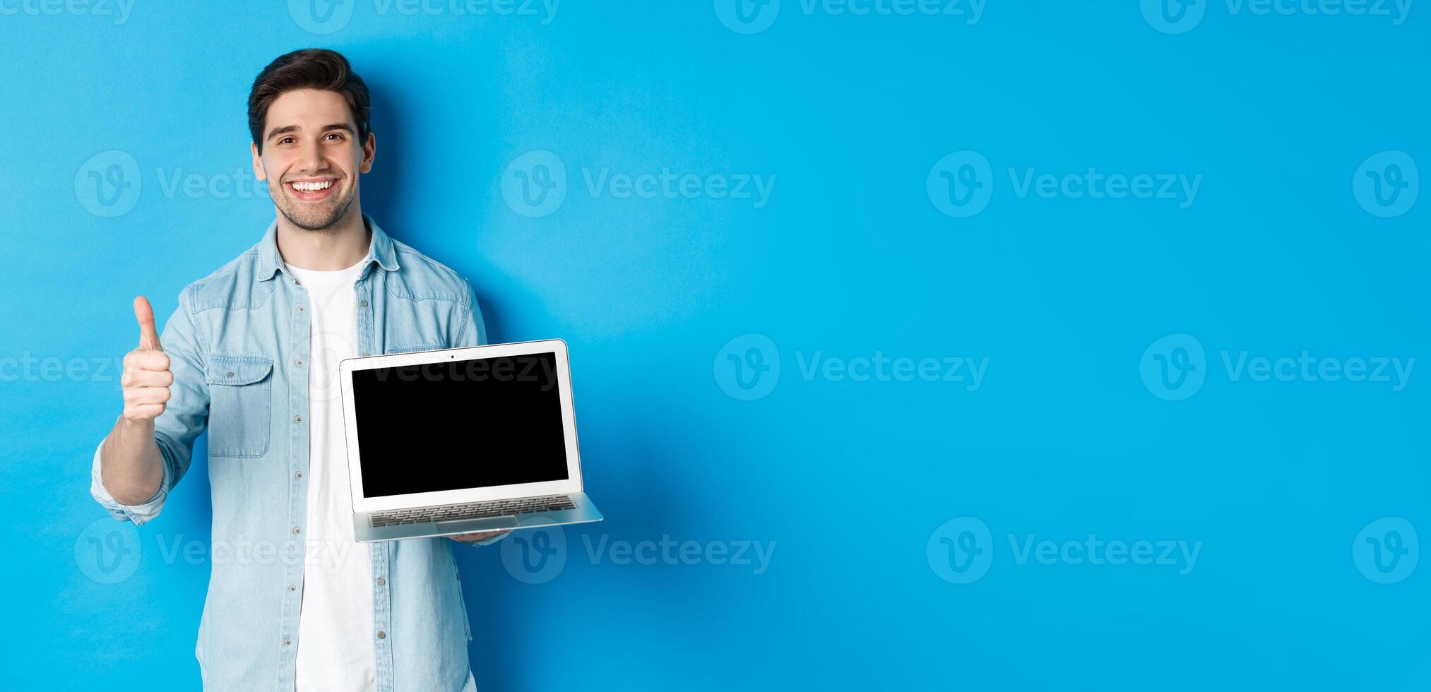 Smiling handsome man showing laptop screen and thumb-up, like promo offer, recommending website, standing against blue background photo