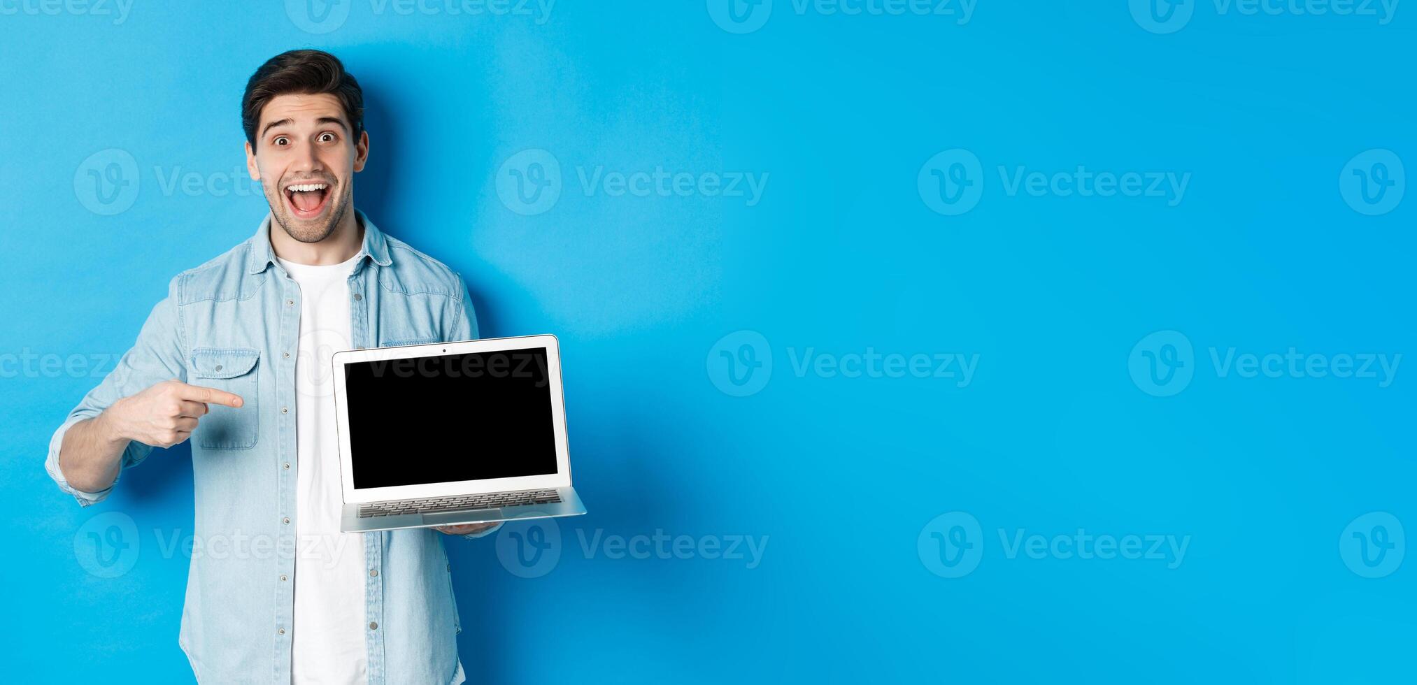 Excited smiling man pointing at laptop screen, showing internet promo offer or website advertisement, standing over blue background photo