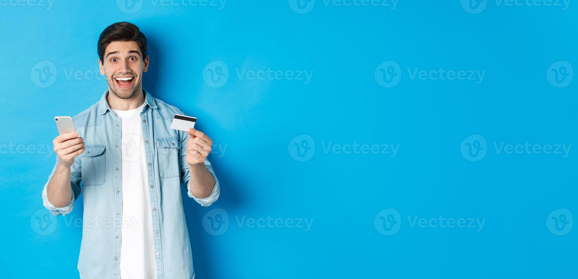 Amazed handsome man shopping online, holding mobile phone and credit card, smiling while paying for internet purchase, standing over blue background photo
