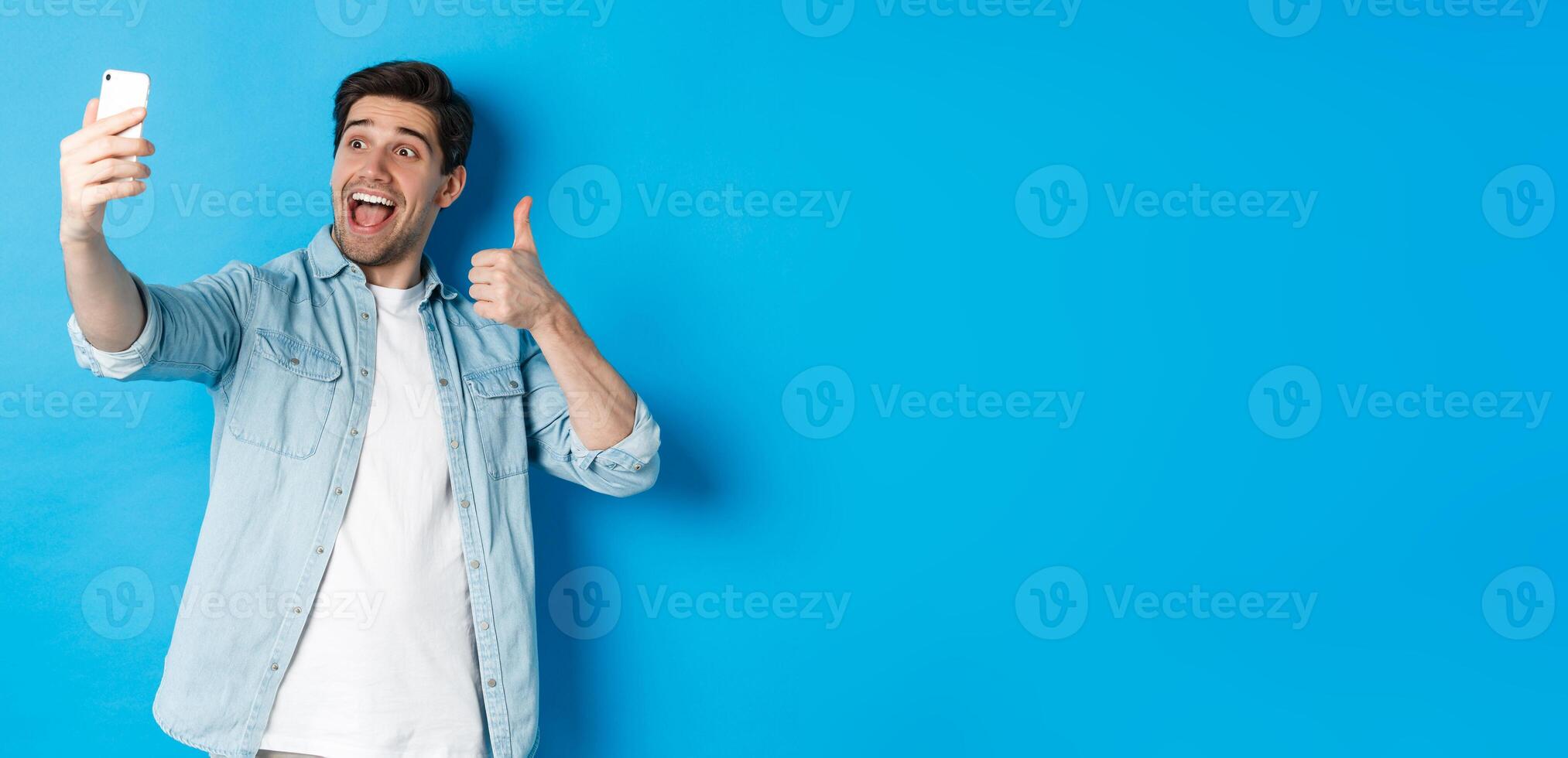 Happy man taking selfie and showing thumb up in approval on blue background, holding mobile phone photo