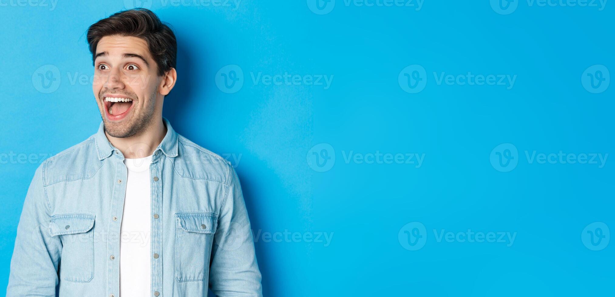 Close-up of handsome caucasian male model looking amazed left, checking out advertisement, standing over blue background photo
