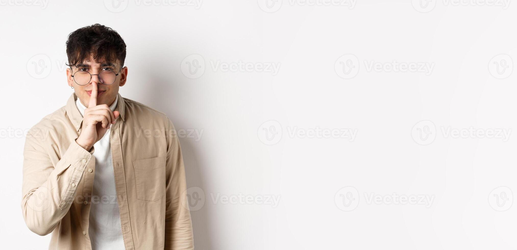 Cheerful handsome man in glasses share a secret, making surprise, lean at camera with hush gesture and gentle smile, telling to be quiet, show taboo sign, white background photo