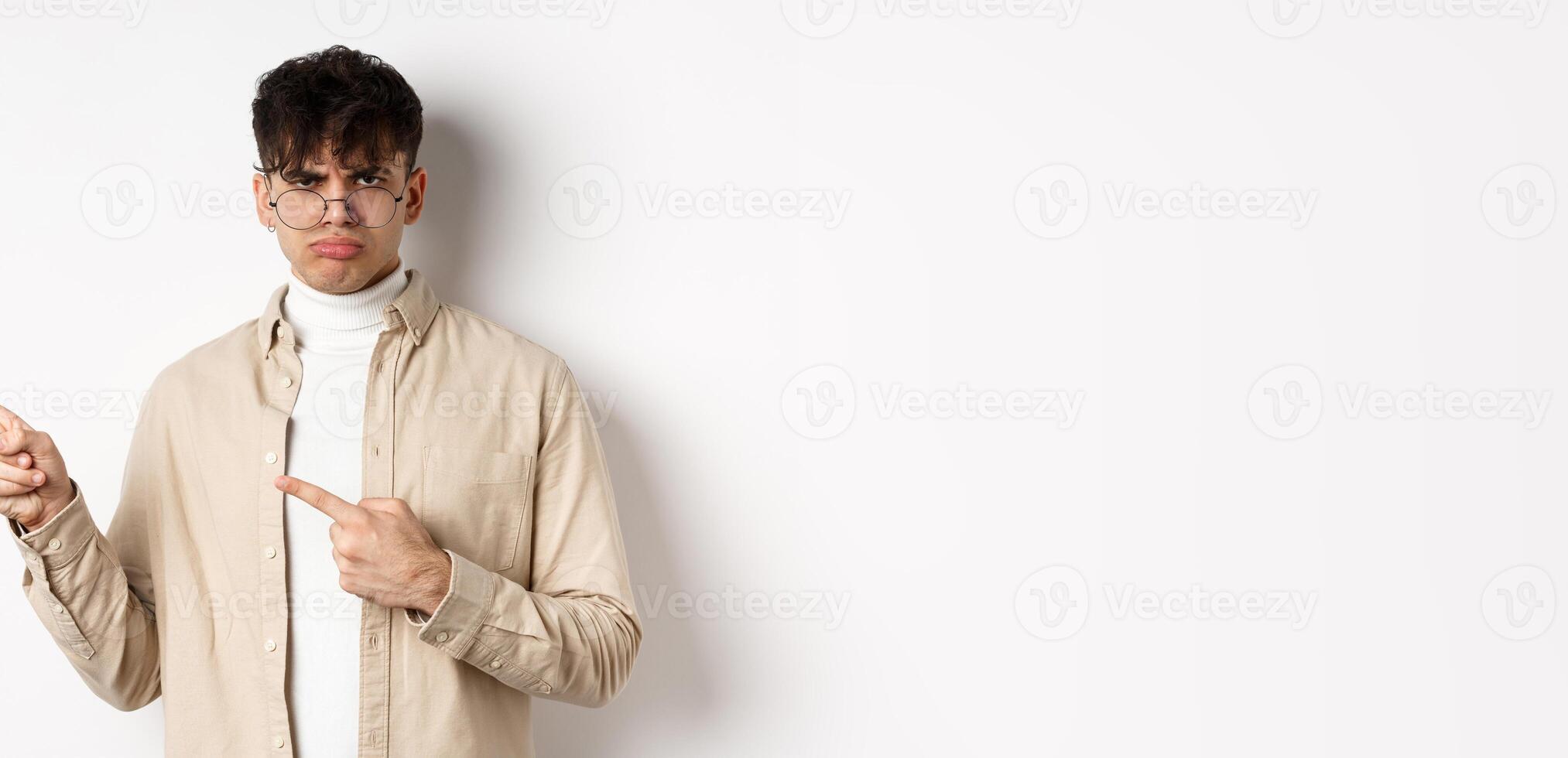 Sad and grumpy boy pointing fingers left, sulking with childish expression, complaining on something bad, standing on white background photo