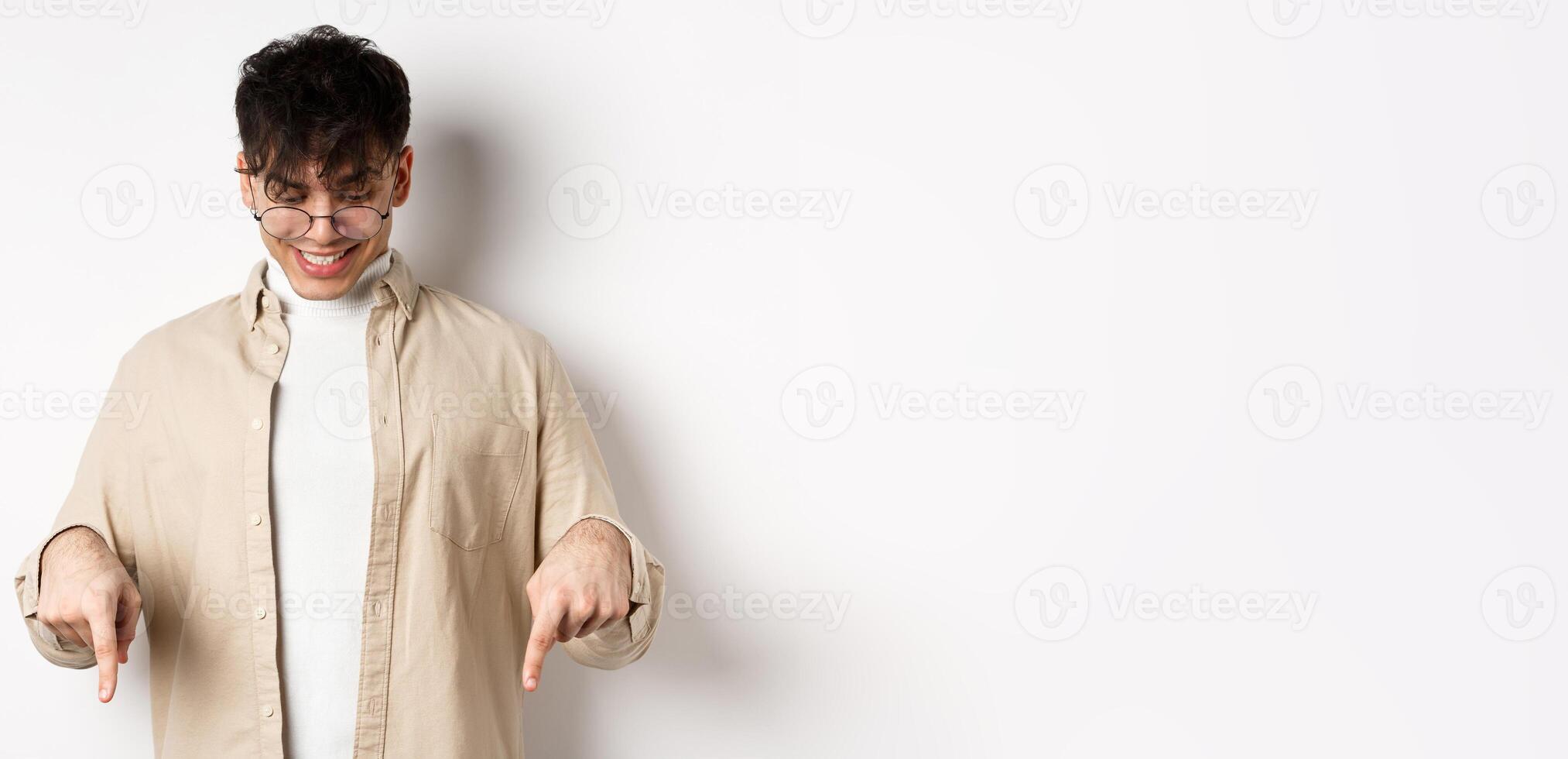 Stylish modern guy in glasses pointing, looking down with happy smile, showing banner, standing on white background photo