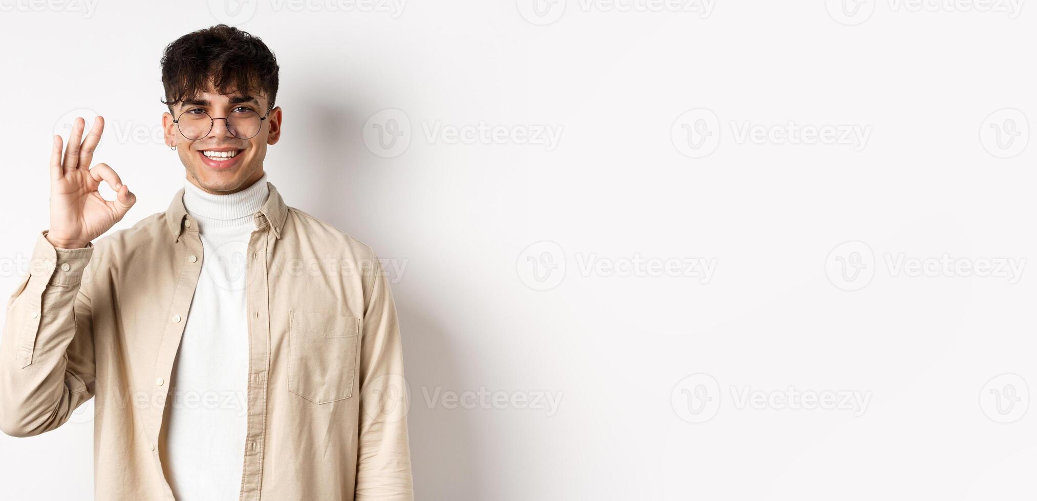 Image of handsome european man in glasses showing okay gesture, smiling satisfied, praise good quality, standing on white background photo