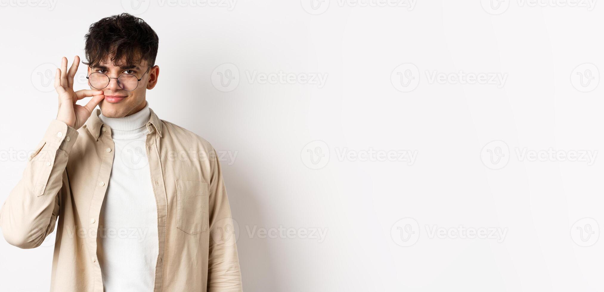 Smiling cunning guy in glasses shut his mouth on zipper, showing seal gesture near lips and smiling devious, keeping a secret, standing on white background photo