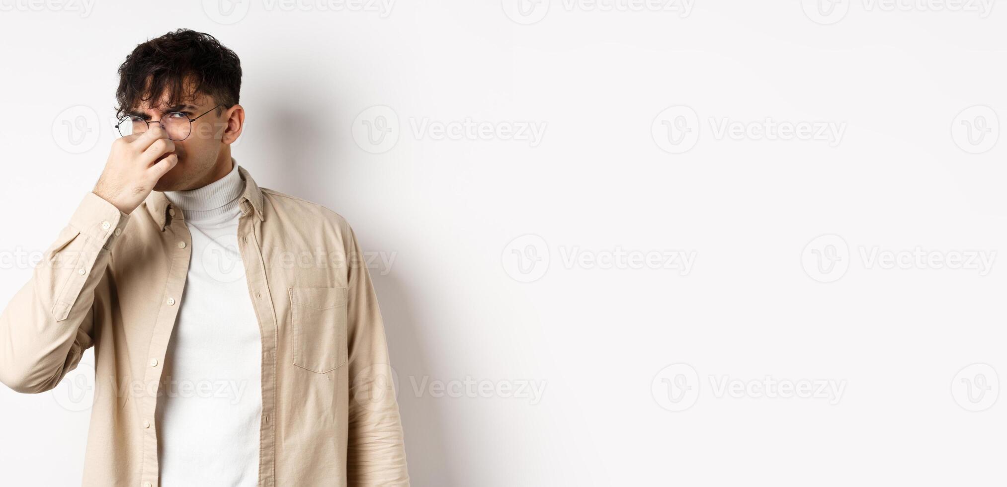 Image of disgusted guy shut his nose from awful smell, looking at something disgusting and stinky, standing on white background photo